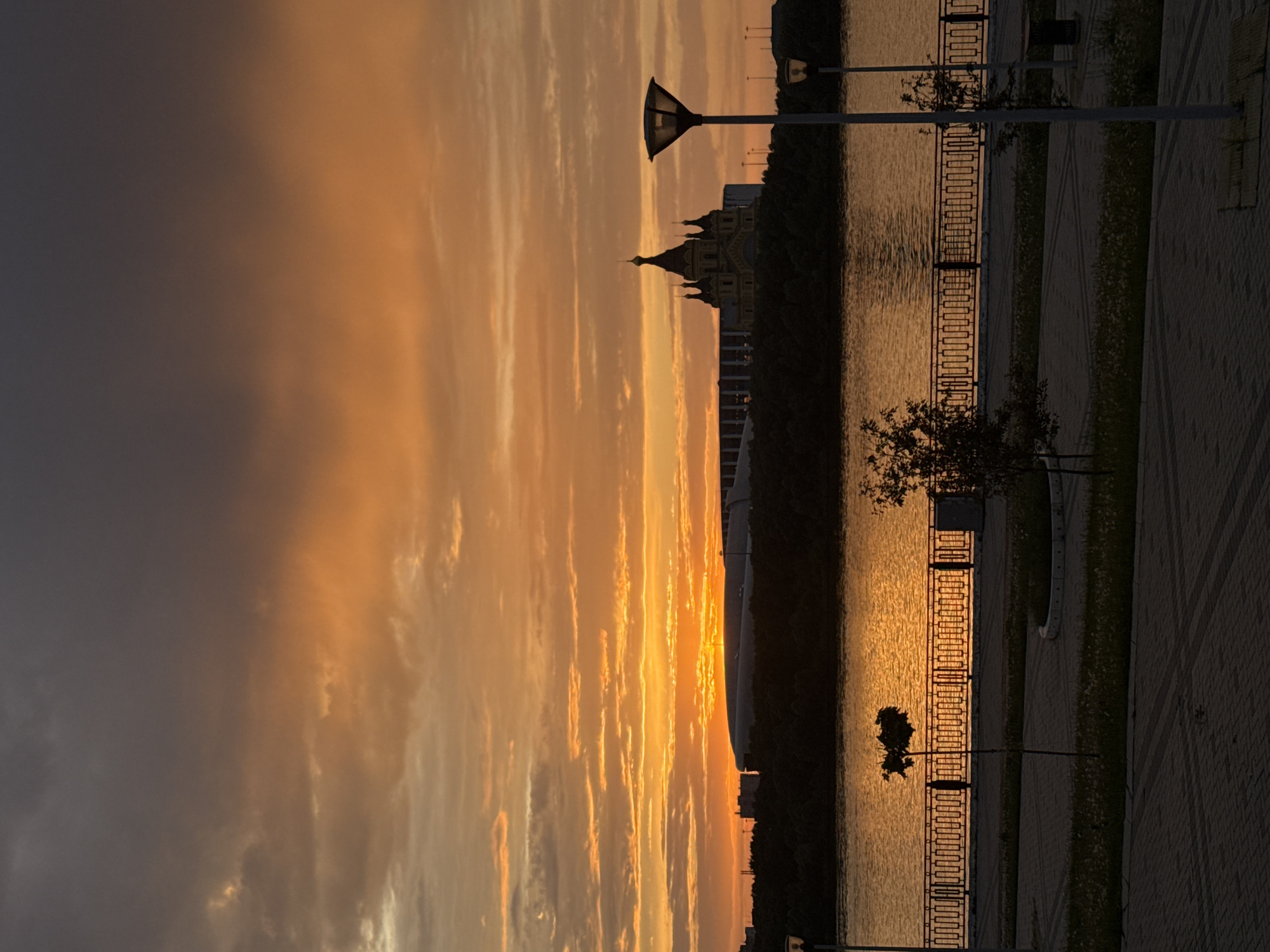 The image depicts a sunset view of Nizhny Novgorod, Russia. The sky is filled with orange and gray clouds. The reflection of the setting sun is visible on the water's surface. A church and other buildings are silhouetted against the bright sky. A waterfront promenade with streetlights, a fence, trees, and paving stones runs along the water.