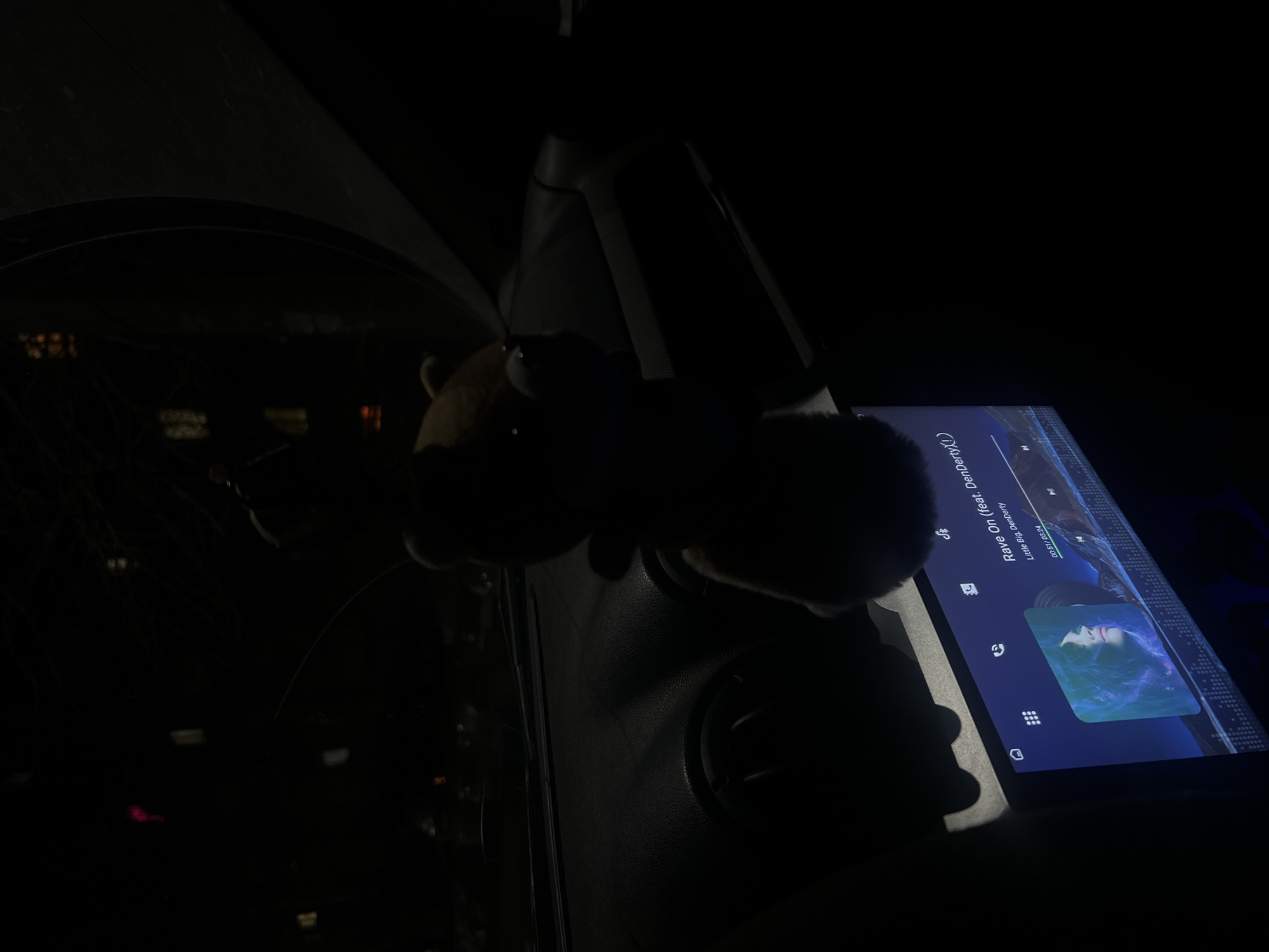 The image captures the interior of a vehicle at night, likely in Chaikovsky, Russia, given the user's location. A stuffed animal rests on the dashboard near an integrated infotainment screen. The screen displays the track "Rave On" by Little Big featuring DenDerty, paused at 0:51 of a 3:24 runtime. The dashboard, vents, and ambient light contribute to a predominantly dark and shadowy composition. Outside the car window, blurred city lights are faintly visible.