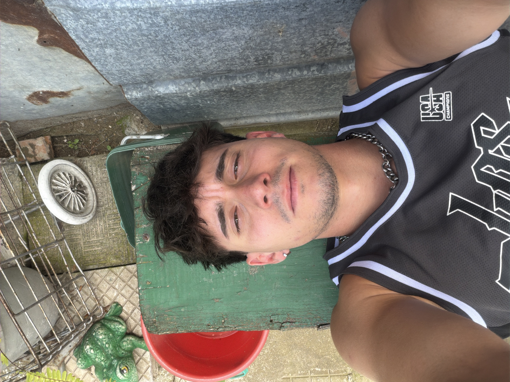 An overhead selfie captures a young adult male lying outdoors in Krumovo, Bulgaria. The man, appearing to be in his 20s, has dark, disheveled hair, a light mustache, and stubble, with a silver hoop earring in his left ear. He wears a dark grey sleeveless basketball-style jersey with white and light grey stripes along the collar and armholes, a silver chain necklace, and a white "USA CHAMPIONS" logo. His gaze is directed at the camera with a neutral expression. He is positioned against a weathered green wooden surface. To the left, on a concrete and tiled ground, are a partially visible red plastic bucket, a green frog statue, a metal cage-like object, and a white spoked wheel. To the right, a corrugated metal sheet with rust stains stands vertically, bordered by dirt ground with sparse vegetation. The scene is brightly lit by natural light.