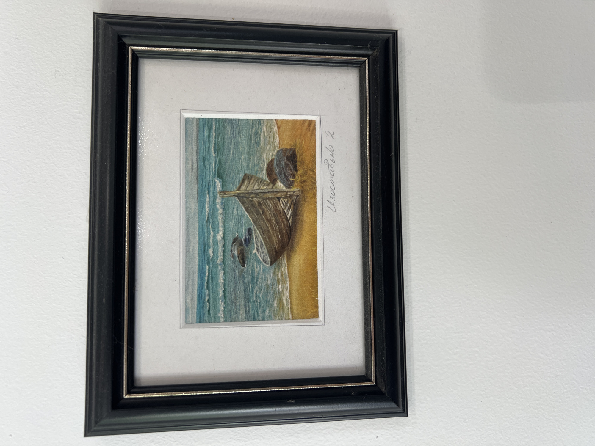 A framed painting is mounted on a white wall. The frame is black with a thin silver inner border. The painting depicts a shipwreck on a beach next to the ocean. The artist's name "Изгостаева 2" is written in light gray cursive along the right edge of the painting. The location is Unknown, Unknown.