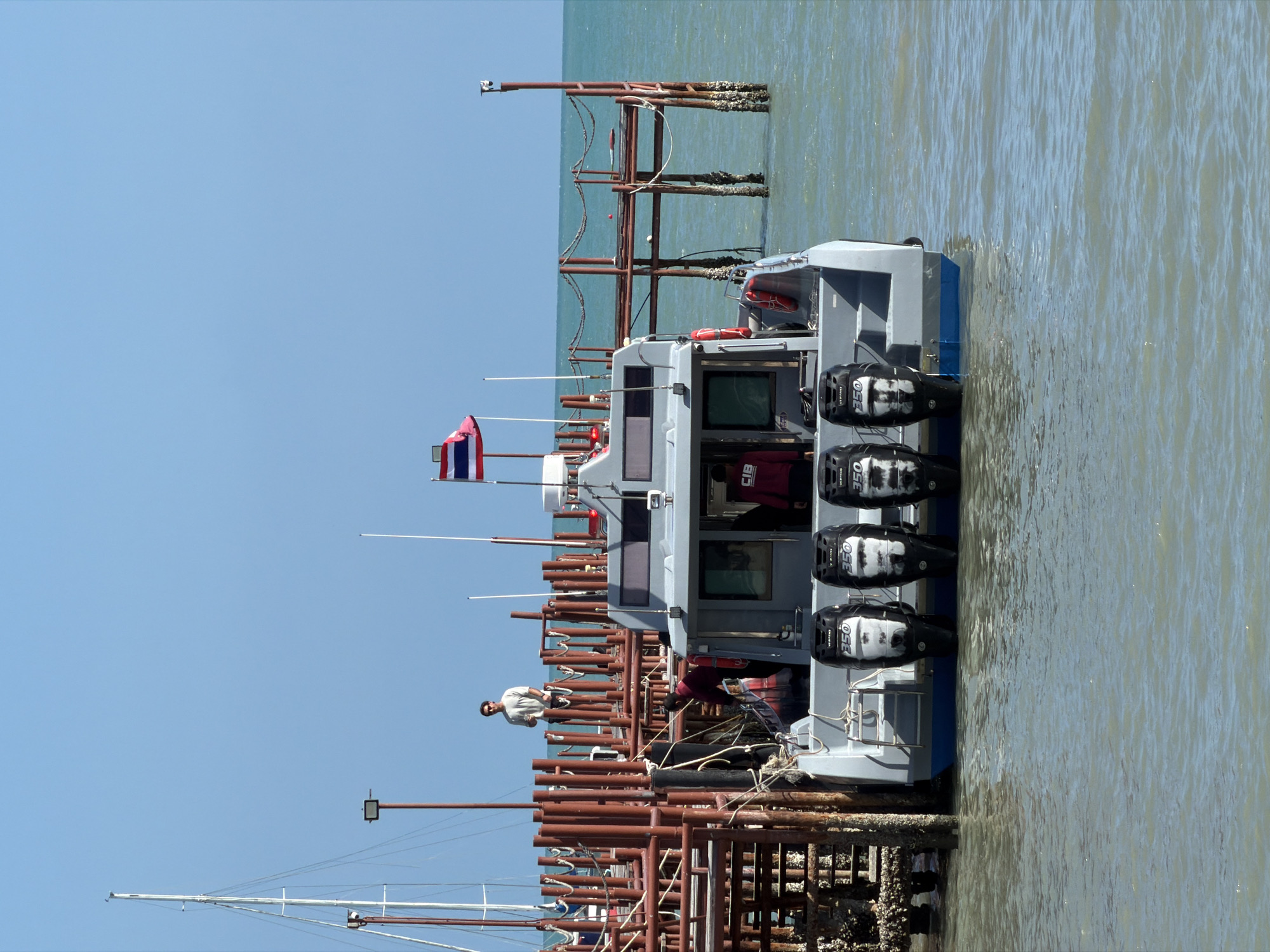 A commercial boat with three outboard motors is docked at a pier in the district of Ko Samui, Thailand. The boat is gray and blue with a white cabin. A Thai flag is visible on the boat's mast. Several people are on the boat and on the pier. The water is choppy and the sky is clear. The pier is made of wood and metal. There are several other boats in the background.