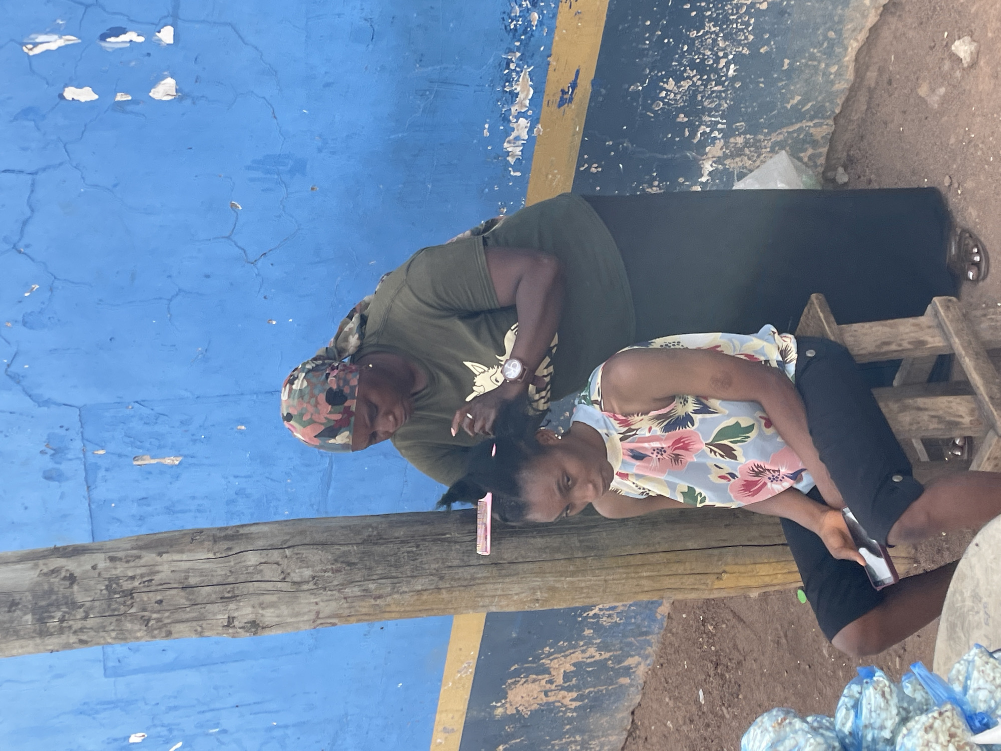 In an outdoor setting in Jos, Nigeria, an adult woman wearing a camouflage headscarf, a green T-shirt, and a watch on her left wrist stands and styles the hair of a young girl sitting on a wooden stool. The girl is wearing a floral-patterned top and dark shorts and holds a bottle in her right hand. A pink comb is stuck in her hair. The background is a blue wall with a yellow stripe, showing signs of wear and tear. A weathered wooden beam runs horizontally in the lower half of the image. Bags of popcorn are visible at the lower right corner. The ground appears to be dirt or gravel.