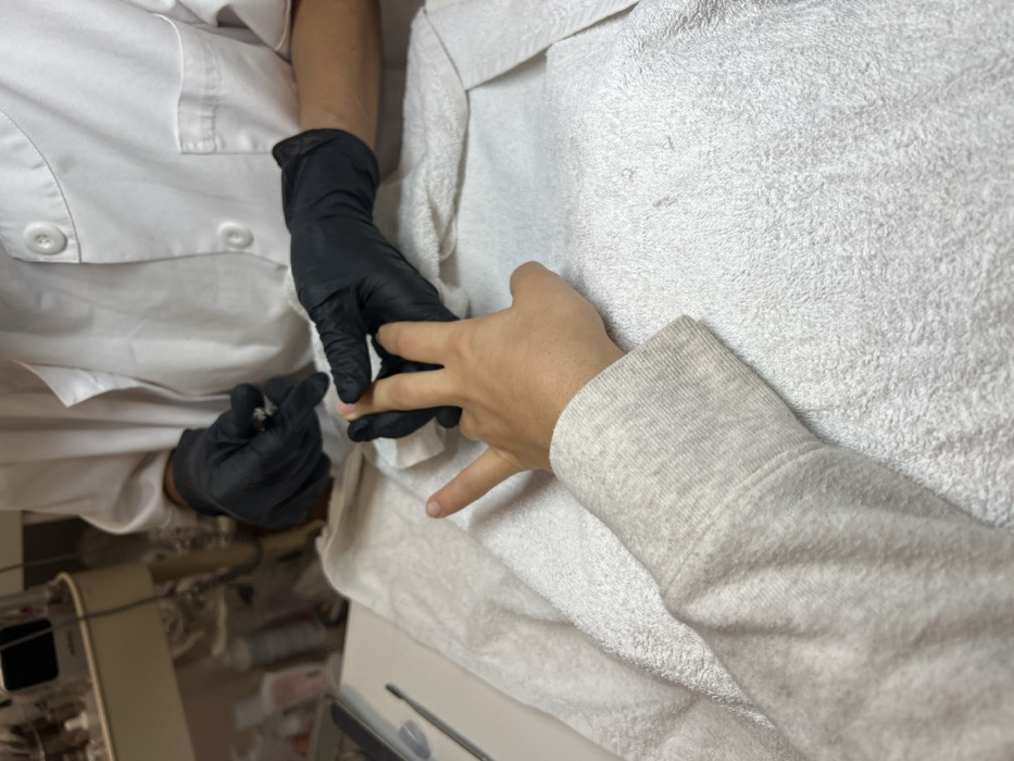 The image depicts an indoor scene in Lordelo do Ouro e Massarelos, Portugal, focusing on a manicure service. A person's right forearm, clad in a light grey long-sleeved garment, and their hand rest on a white terrycloth towel that covers a table surface. A second individual, wearing a white uniform jacket with two visible black buttons and black nitrile gloves, is performing a nail treatment. The gloved hands of the second person are actively holding the client's index and middle fingers. A silver-colored nail tool, consistent with cuticle nippers, is visible in the manicurist's left gloved hand. The background is softly blurred, revealing shelves or a trolley with multiple small bottles and containers, indicative of a professional nail salon setting.