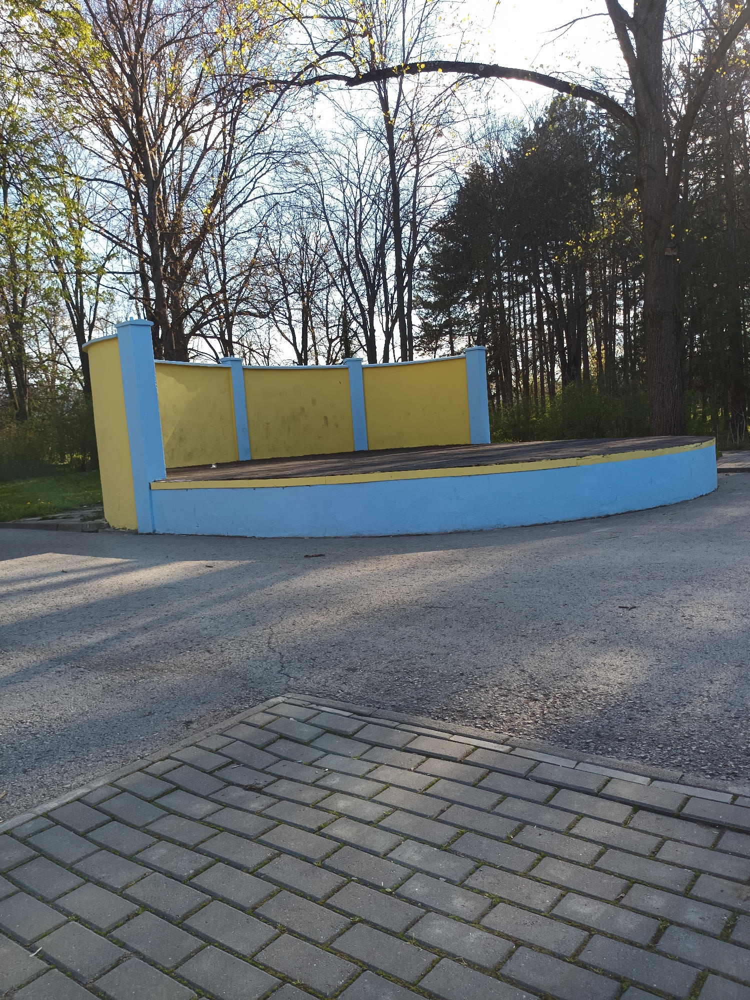 The image depicts a small, outdoor stage in a park-like setting. The stage is painted yellow and blue and has a curved front. The stage flooring appears to be made of wood. The ground in front of the stage is paved with asphalt, with a section of gray brick paving in the foreground. Background includes bare trees on the left and taller evergreen trees on the right. The location is in Targovishte, Bulgaria. There are no people visible in the image.