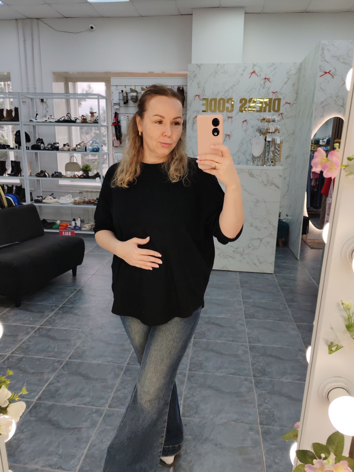 A middle-aged woman with blonde curly hair is taking a selfie in what appears to be a retail store in Chaikovsky, Russia. She is wearing a loose-fitting black top, blue jeans, and gold hoop earrings. She is holding a pink smartphone in her right hand and has her left hand resting on her stomach.
In the background, there is a display of shoes on shelves, a black loveseat, a wall with the words "DRESS CODE" written backward, and a mirror with lights. The floor is tiled with gray square tiles.