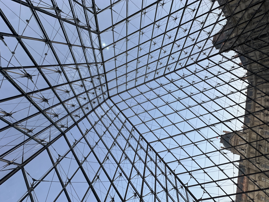 The image presents an upward view from inside or directly beneath the Louvre Pyramid in Paris, France. The dominant feature is the intricate geometric framework of the pyramid, consisting of numerous dark metal beams intersecting to form a grid of diamond-shaped glass panes. Through the transparent glass, a clear, light blue sky is visible, with a small, bright sunburst apparent in the upper right quadrant. Below the pyramid's structure, sections of the classical stone buildings of the Louvre Palace are discernible, displaying multiple windows, intricate architectural details, and mansard roofs. No individuals, specific actions, or interactions are depicted within the scene.