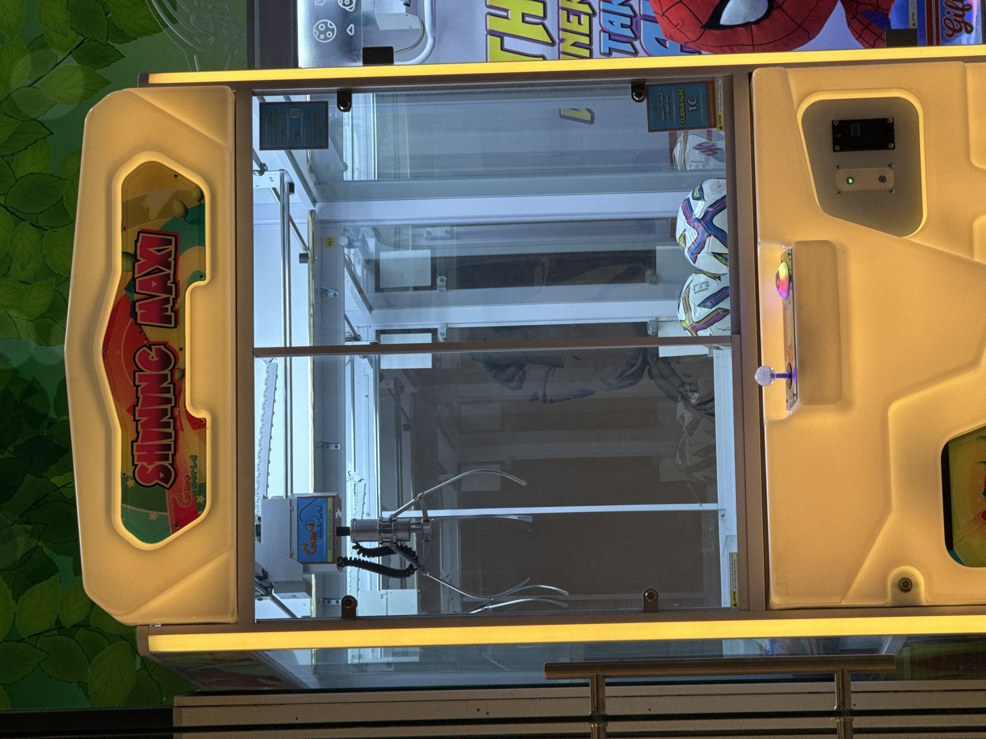 The image shows a crane game machine in an indoor location. The machine is brightly lit with yellow lights, and the cabinet is decorated with text and graphics, including "SHINING MAXI" and a Spiderman image. The inside of the machine contains several soccer balls. The crane claw is visible in the lower part of the cabinet. A control panel with buttons is located on the side. There are some printed texts which specify the price for a game in Euros and the start of game by inserting coins. Green leaves are seen on the left of the machine which may be a part of decoration of the location. The game is in an unidentifiable city.