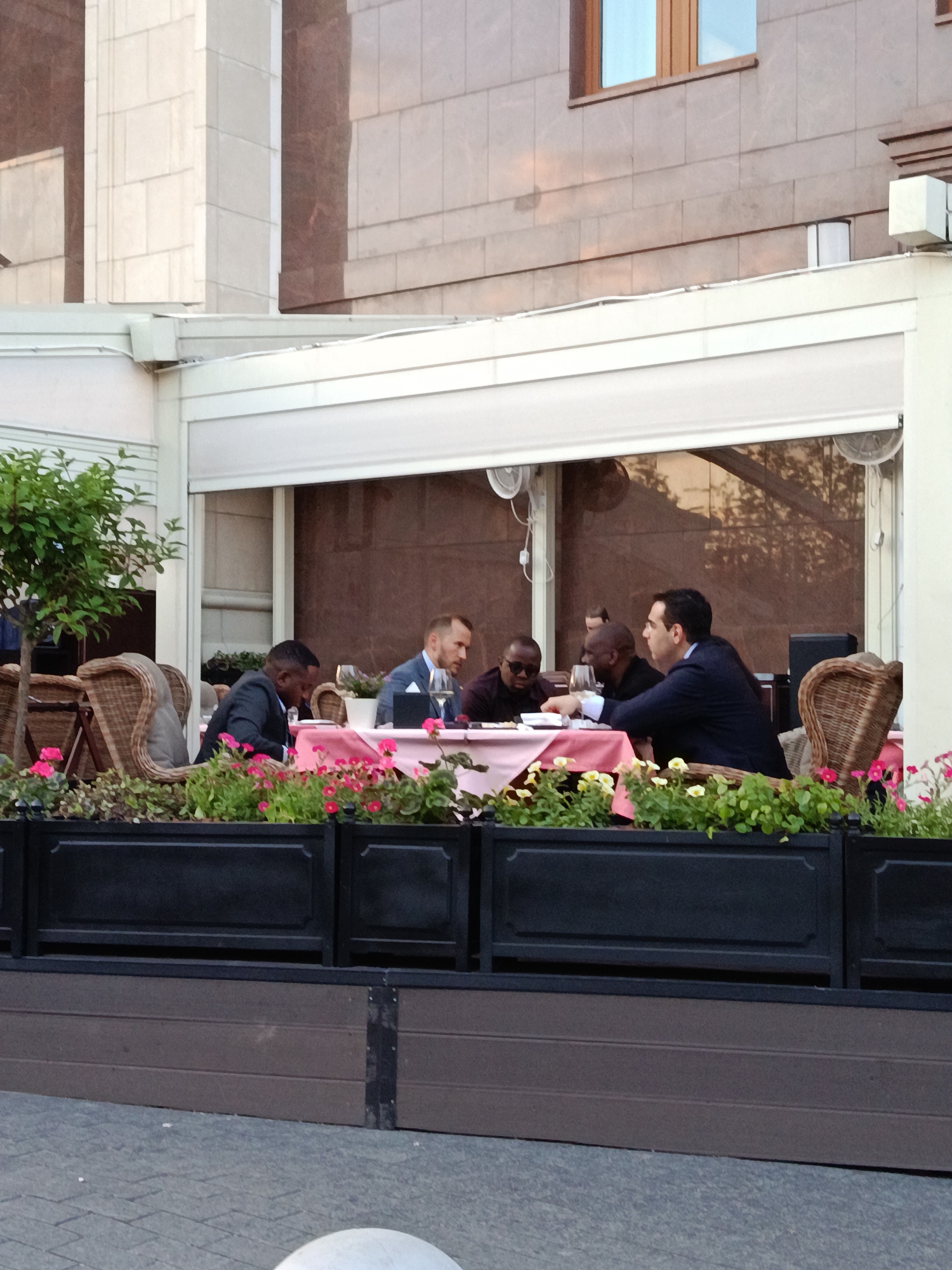 The image depicts a group of men gathered at an outdoor dining area, likely a restaurant or cafe patio. Approximately six men are seated around a round table, which is covered with a pink tablecloth and holds wine glasses and other items. They appear to be engaged in conversation or a business meeting, with some men looking at each other while others look down at the table. One man in a dark suit on the right is reaching towards the table, while another in a blue jacket holds a glass.
The dining area is furnished with wicker chairs and bordered by black planters filled with green foliage and bright pink flowers, possibly petunias. A small potted tree is visible on the left side of the frame. Below the planters, there are brown wooden-like panels, and the foreground shows grey paved ground with part of a white bollard visible at the bottom.
Above the seating area, a white awning or canopy structure extends, beneath which two circular, fan-like objects are suspended. In the background, a building with a facade of light brown or grey stone tiles and a brown-framed window is visible. The soft, ambient lighting suggests the time of day is late afternoon or early evening, indicative of pleasant weather suitable for outdoor dining. No visible text is present in the image.