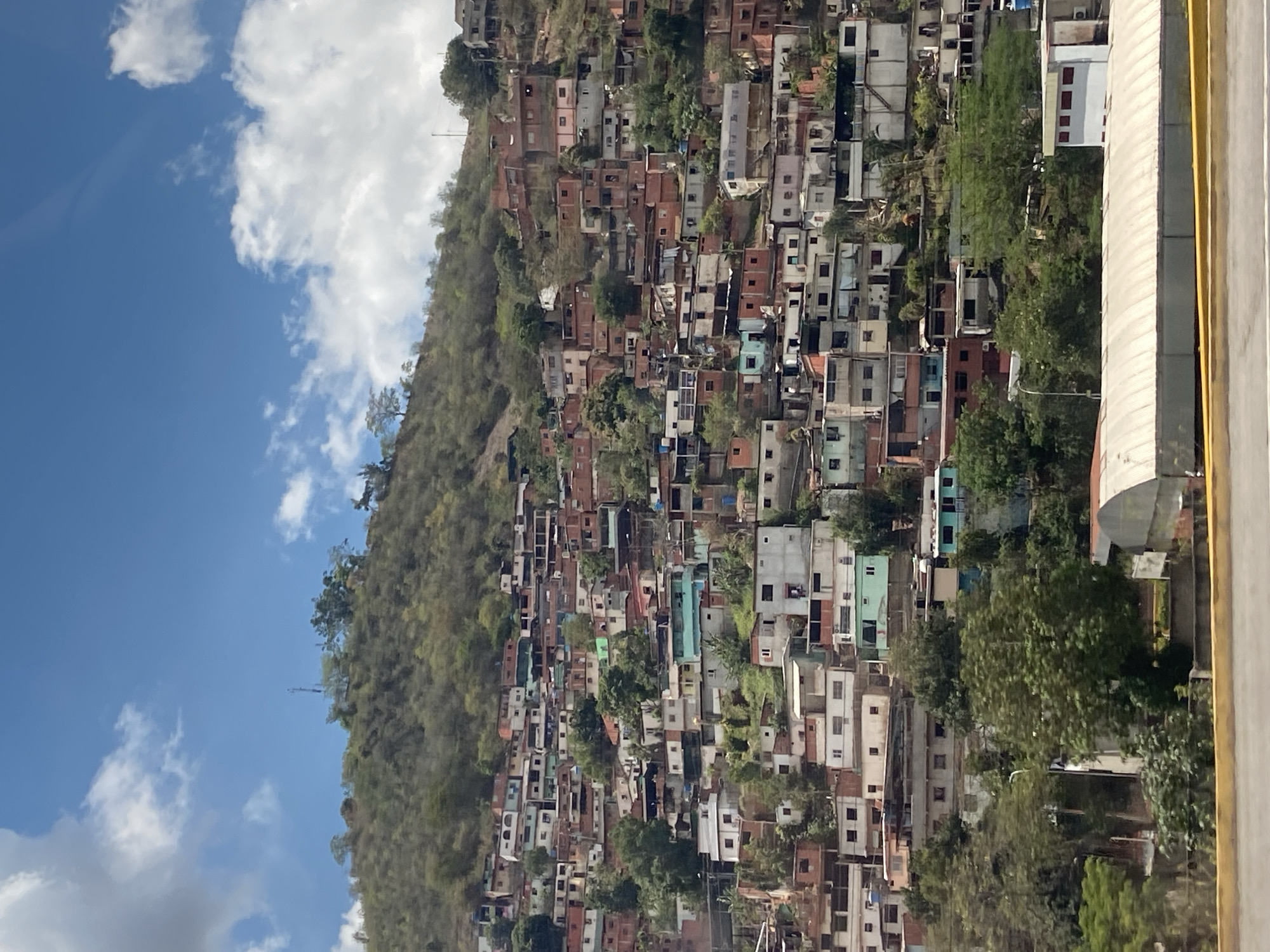 The image displays a densely populated hillside in Guarenas, Venezuela, characterized by informal residential structures. The steep, vegetated slope is covered with numerous multi-story buildings, predominantly constructed from concrete and brick, featuring a mix of flat and corrugated metal roofs. These structures exhibit varied finishes, including unpainted concrete, exposed red brick, and pastel paint colors such as white, light blue, pink, and muted green. They are arranged in a terraced fashion up the hillside. Sparse trees and shrubs are interspersed among the dwellings and on the upper, less dense portions of the hill. Communication towers are visible on the hill's summit. At the base of the hill, partially obscured by trees, are a large, curved metallic-roofed building and a smaller white structure with red-accented windows. The foreground exhibits a yellow-striped concrete barrier, suggesting the viewpoint is from a roadside or elevated platform. The sky above is blue with scattered white cumulus clouds. No discernable individuals are present.