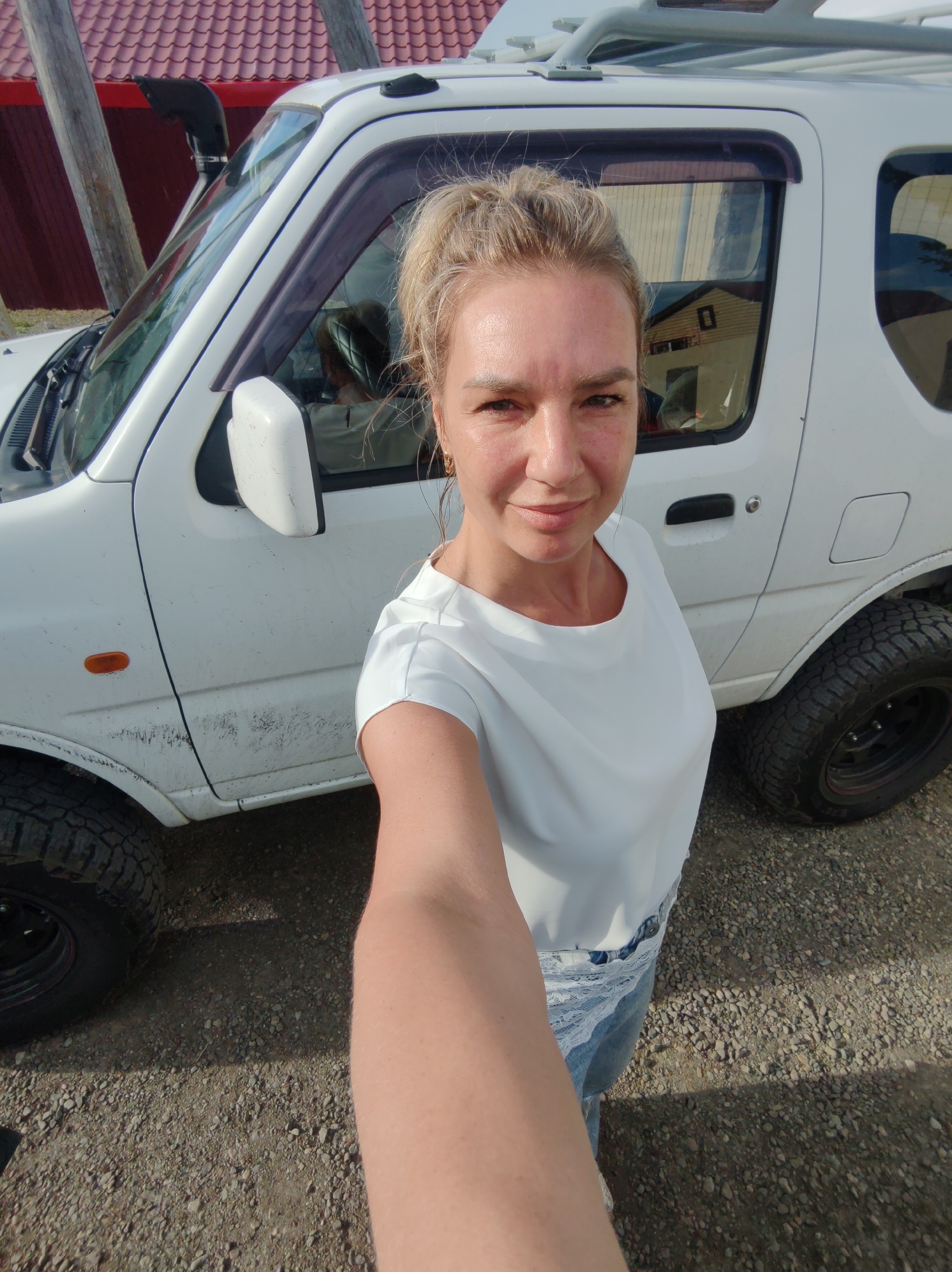 A woman with blonde hair pulled up in a bun, wearing a white t-shirt and denim shorts with white lace trim, takes a selfie in front of a white Suzuki Jimny SUV. The SUV is parked on a gravel surface outside. The vehicle has a snorkel attached to the driver's side windshield pillar, and its tires appear to be off-road tires. The background shows a red-tiled roof and wooden structures, consistent with a rural or suburban setting in Paratunka, Russia. The lighting suggests it is daytime, with some shadows indicating the sun's position.