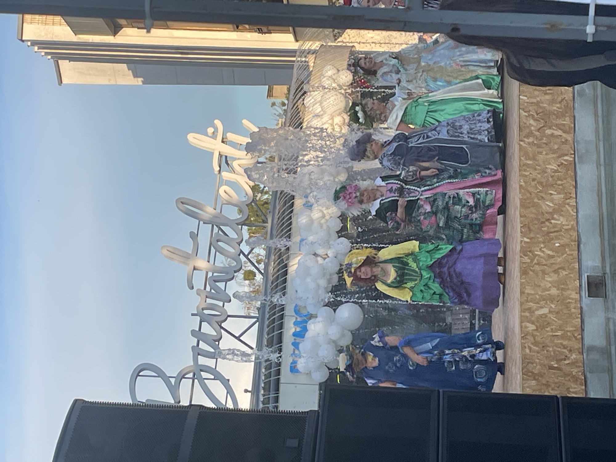 The image captures an outdoor event with several individuals dressed in elaborate historical costumes, likely from the 18th or 19th century, standing on a wooden stage. The costumes feature wide skirts, corsets, and ornate hats. There are fountains with water cascading behind the performers, and white balloon-like structures are suspended above the stage, possibly part of the decoration. In the background, a building with a distinct architectural style, featuring vertical lines and a stepped facade, is visible. A large speaker is positioned to the left of the frame. The sky above is a clear blue with some faint clouds. The overall scene suggests a performance or a historical reenactment taking place in a public space. The mention of Brest, Belarus, in the prompt cannot be visually confirmed from the image alone.