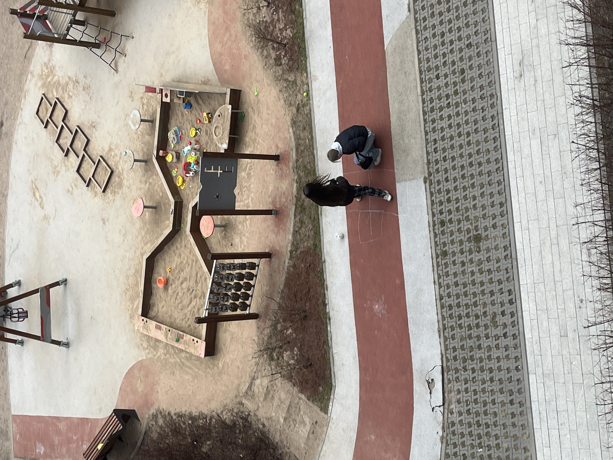 An aerial view captures a playground situated near a pathway. Two individuals are seen near the walkway. To the right of the path, there is a stone-covered ground with shrubs along the right edge. The playground is located to the left of the pathway and features a variety of play structures. Based on the architectural style and urban layout, the image likely depicts a scene in Saint Petersburg, Russia.