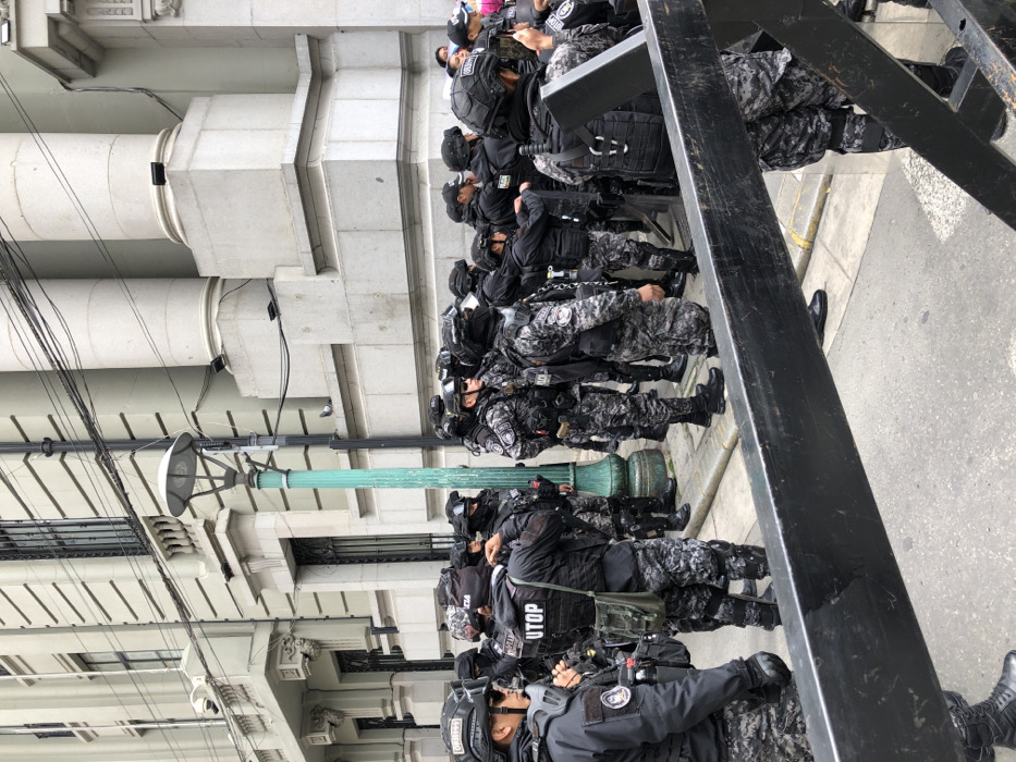 A line of police officers, clad in dark tactical gear with camouflage patterns, stands at rest on a sidewalk next to a building with classical architectural details. They wear helmets and body armor, and some carry batons. A street lamp and metal barriers are visible. The officers appear to be waiting, possibly for deployment in a public order situation. "UTOP" is visible on one officer's gear.