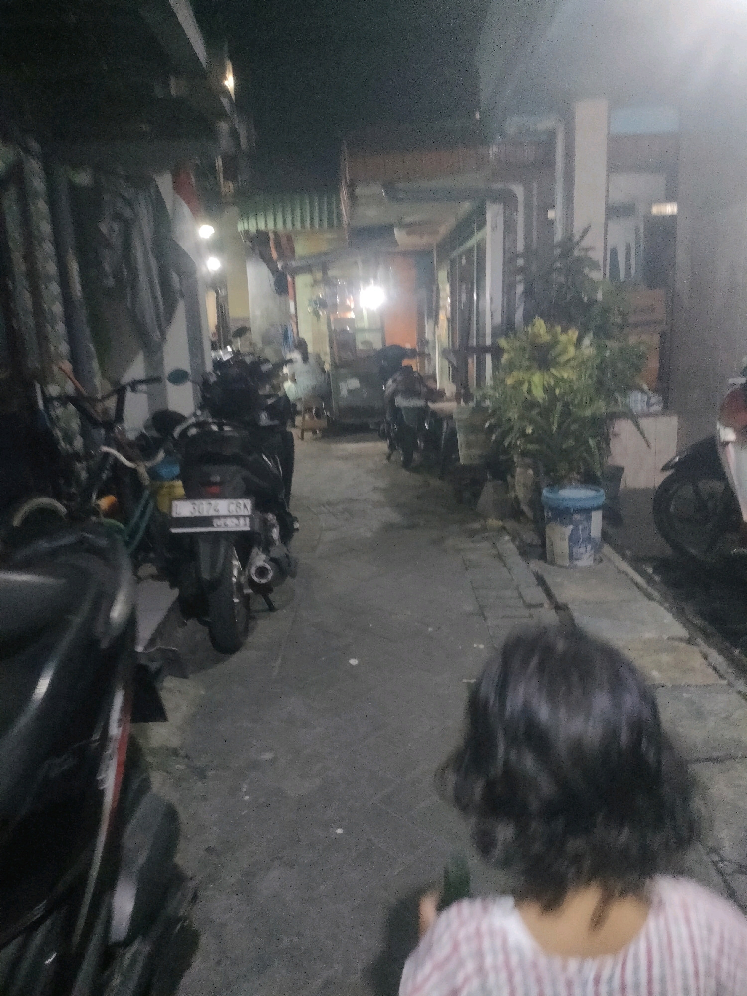 A narrow alleyway in Surabaya, Indonesia, is depicted at night. The alley is lined with buildings and features several parked motorcycles. A child with dark, wavy hair is visible in the foreground, facing away from the camera. The child is wearing a striped shirt. The environment is dimly lit, with artificial lights illuminating parts of the alley and casting shadows. The overall impression is of a residential or commercial backstreet scene in a tropical urban setting.