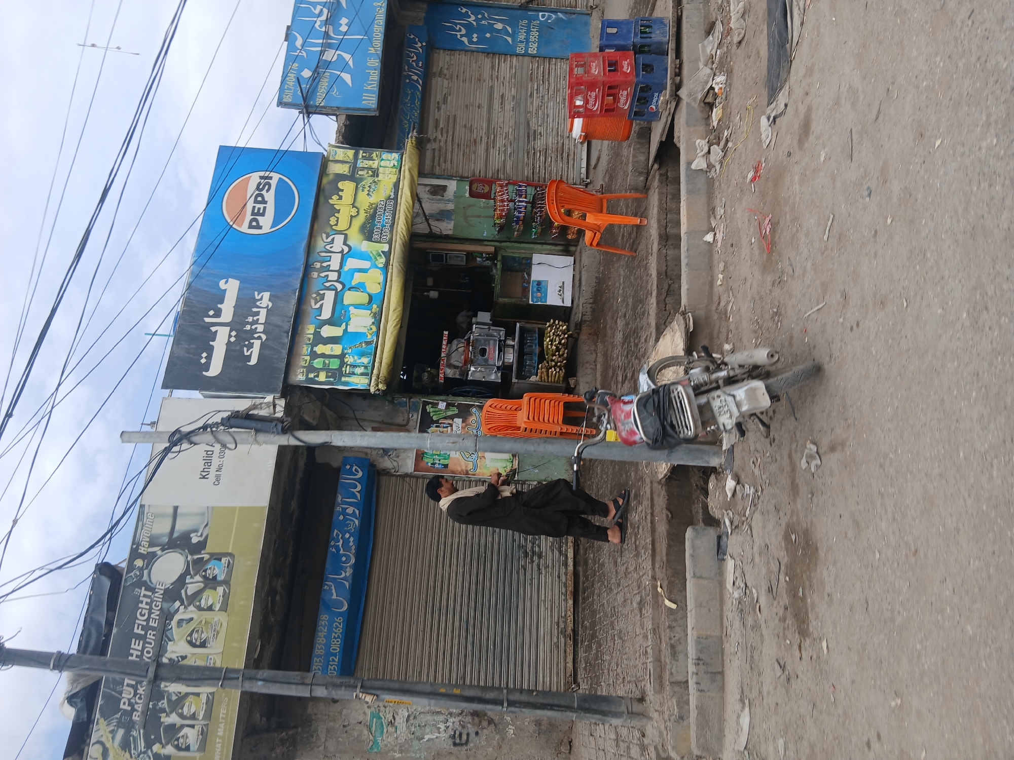 The image depicts a street scene in Quetta City Tehsil, Pakistan. Several businesses are visible, including a shop with a large Pepsi sign and a store selling Havoline engine oil. A man wearing traditional Pakistani clothing stands in front of a closed store, with a motorcycle parked nearby. The street appears somewhat cluttered with discarded items. The sky is overcast. The storefronts have a mix of Urdu and English signage.