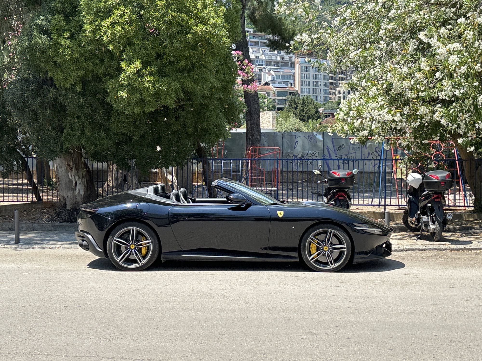 A black convertible Ferrari is parked on an asphalt street in front of a black metal fence. Behind the fence, a large tree with green leaves is visible, and further in the background are buildings and more trees with white flowers. Two motorcycles are parked behind the fence. The street is paved with asphalt, and the weather appears to be sunny.