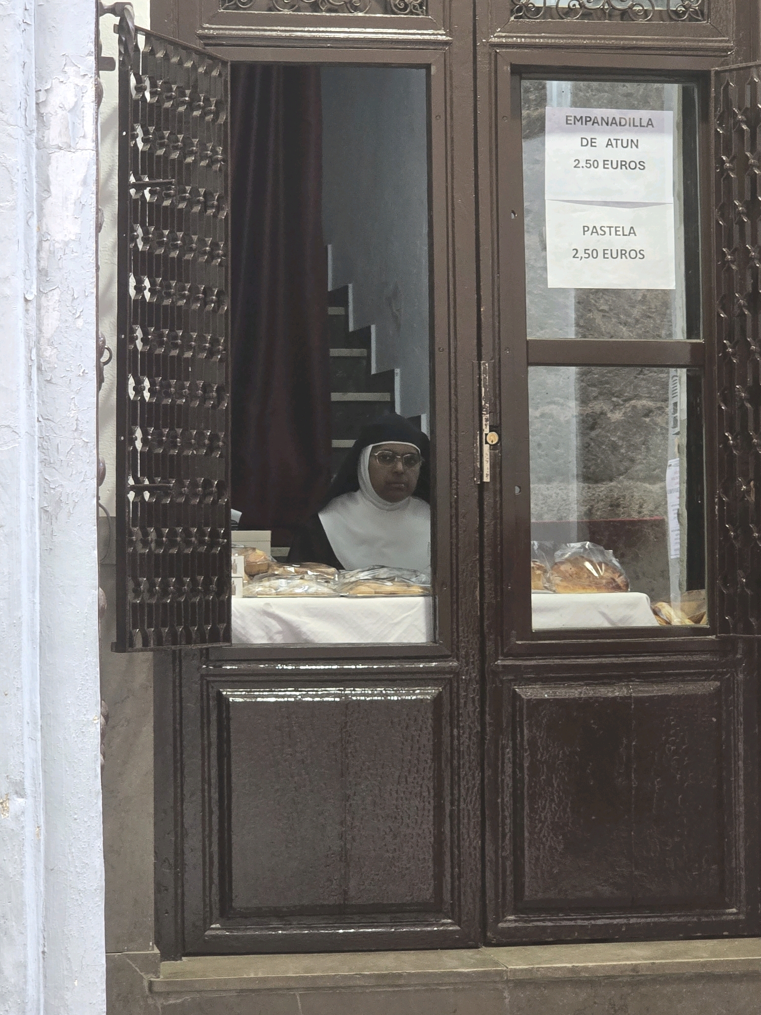 A nun, wearing a traditional habit with a white wimple and black veil, looks out from behind a counter in a shop. The shop appears to be a bakery or food stall. Behind her, a staircase with white steps is visible. On the counter, there are several baked goods, some of which are wrapped in plastic. A sign on the window lists "EMPANADILLA DE ATUN 2.50 EUROS" and "PASTELA 2.50 EUROS." The shop has a dark brown wooden door and window frame with decorative metal grilles on the left side. The exterior wall to the left is painted a pale blue and shows signs of peeling paint. This scene is likely in Granada, Spain, given the Spanish text on the sign and the architectural style.