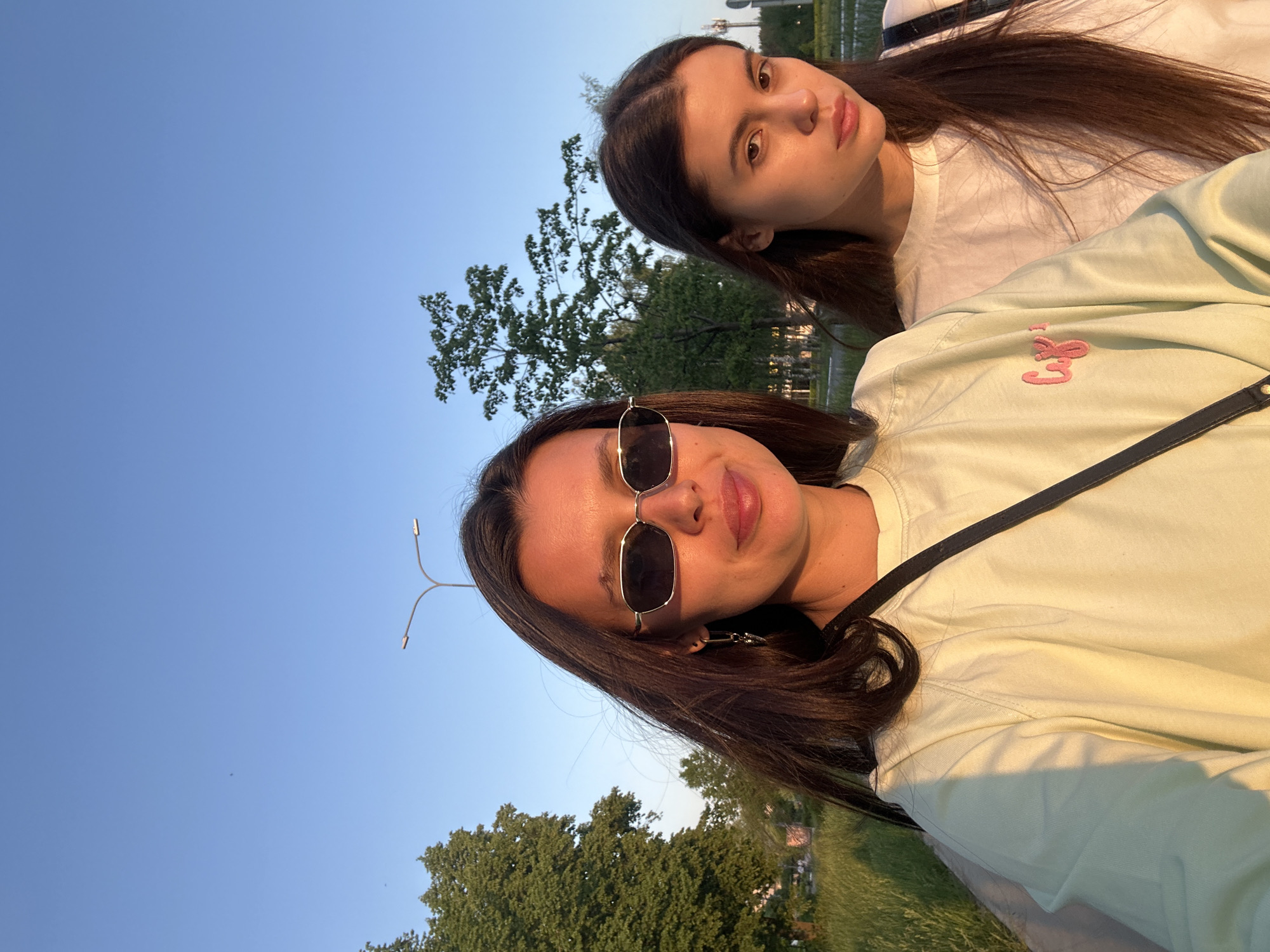 Two young women are in the frame. One woman is wearing sunglasses, a light green sweatshirt, and earrings. The other woman is wearing a white shirt. The location is Chaikovsky, Russia, as indicated by the distinctive street lighting visible in the background. Trees are visible in the background and the sky is a clear blue. The photo appears to be taken outdoors during the daytime.