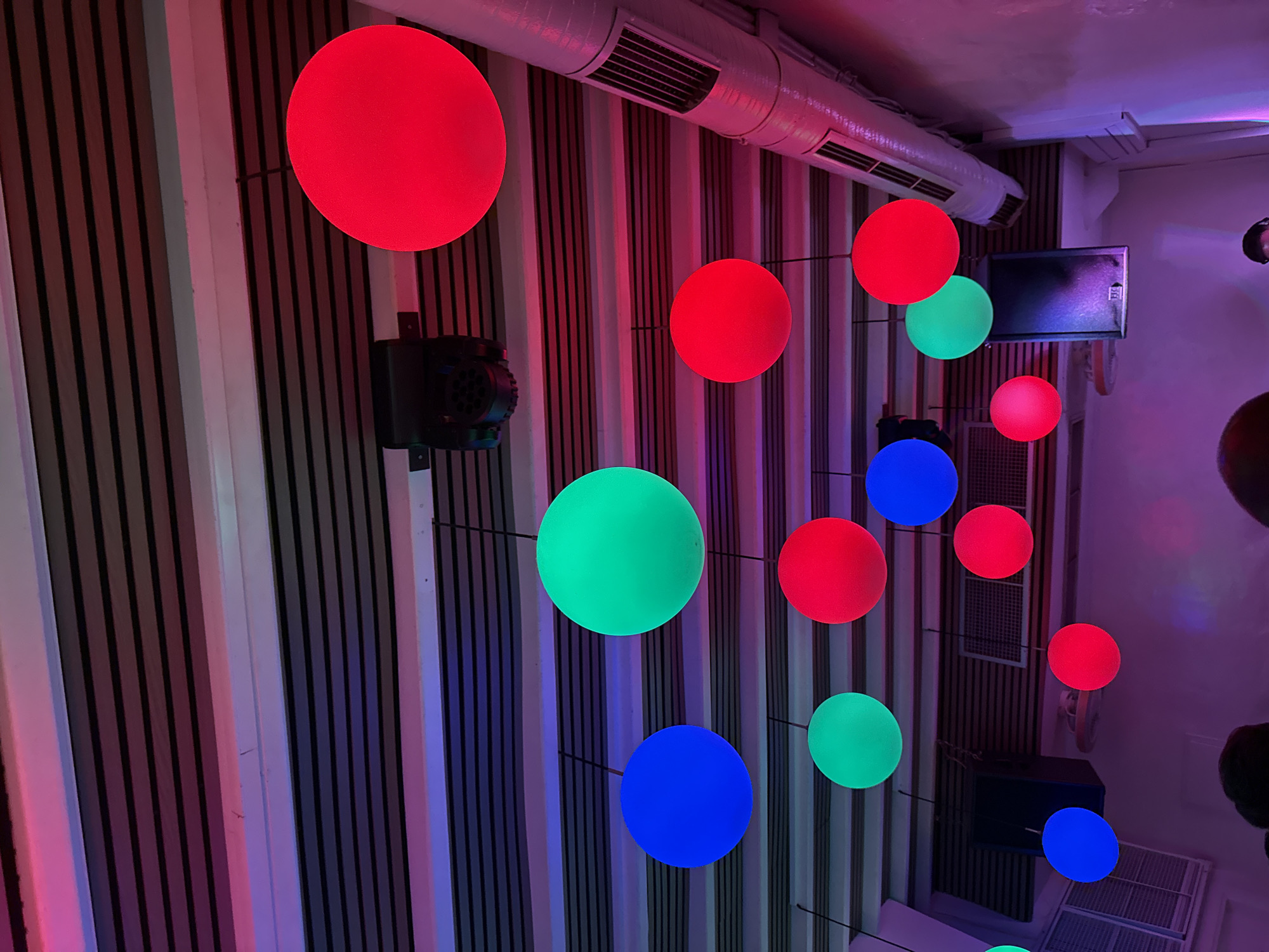 A dimly lit indoor space, likely a club or bar in Mykonos, Greece, is illuminated by numerous colorful, spherical lights suspended from the ceiling. The ceiling itself is constructed with horizontal wooden slats interspersed with darker elements, creating a striped effect. Above, a black stage light fixture is mounted. Various spherical lights, in shades of red, blue, and green, hang at different heights, casting vibrant hues across the scene. A large silver ventilation pipe runs along the right side of the ceiling. In the background, dark speakers are visible. The lower portion of the image shows the tops of people's heads, indicating a gathering. The overall atmosphere is festive and vibrant, characteristic of a nightlife venue.