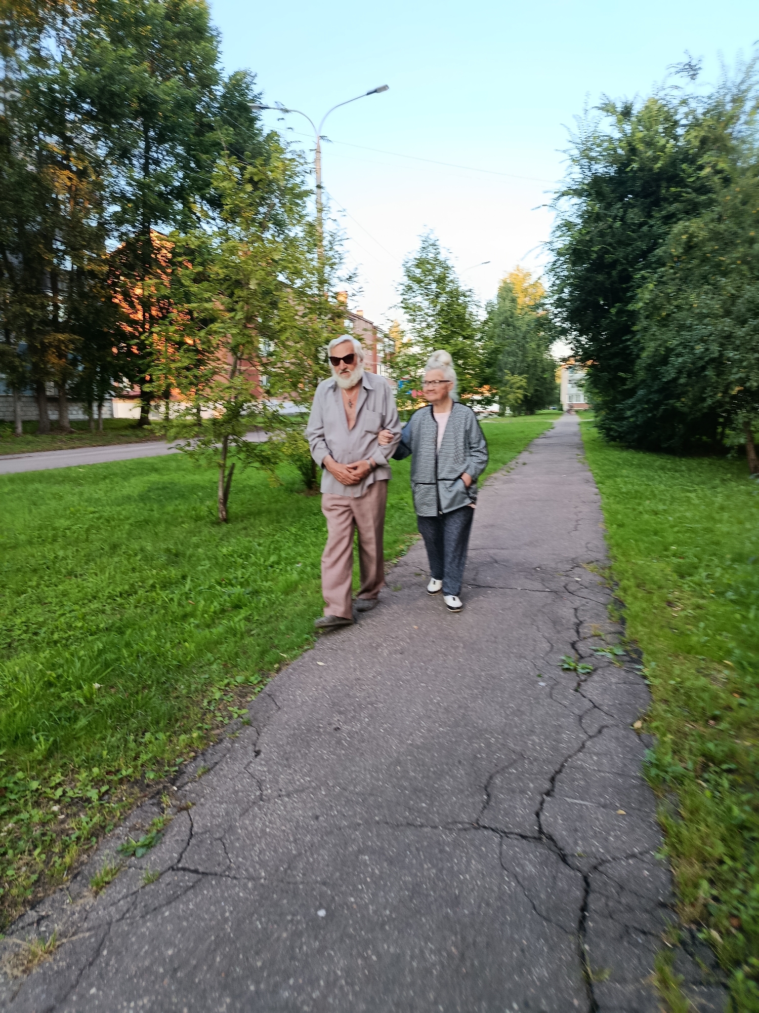 An elderly couple walks down a cracked asphalt path in a park or residential area. The man, on the left, wears a light-colored open-collared shirt, tan pants, and sunglasses. The woman, on the right, wears a striped jacket over a pink top and dark pants, with her hair pulled back. They are walking towards the viewer, with trees and buildings in the background. The scene suggests a leisurely stroll in a moderately urban or suburban environment, possibly in Verea, Russia, given the architecture and vegetation. The lighting indicates it is likely late afternoon or early evening.