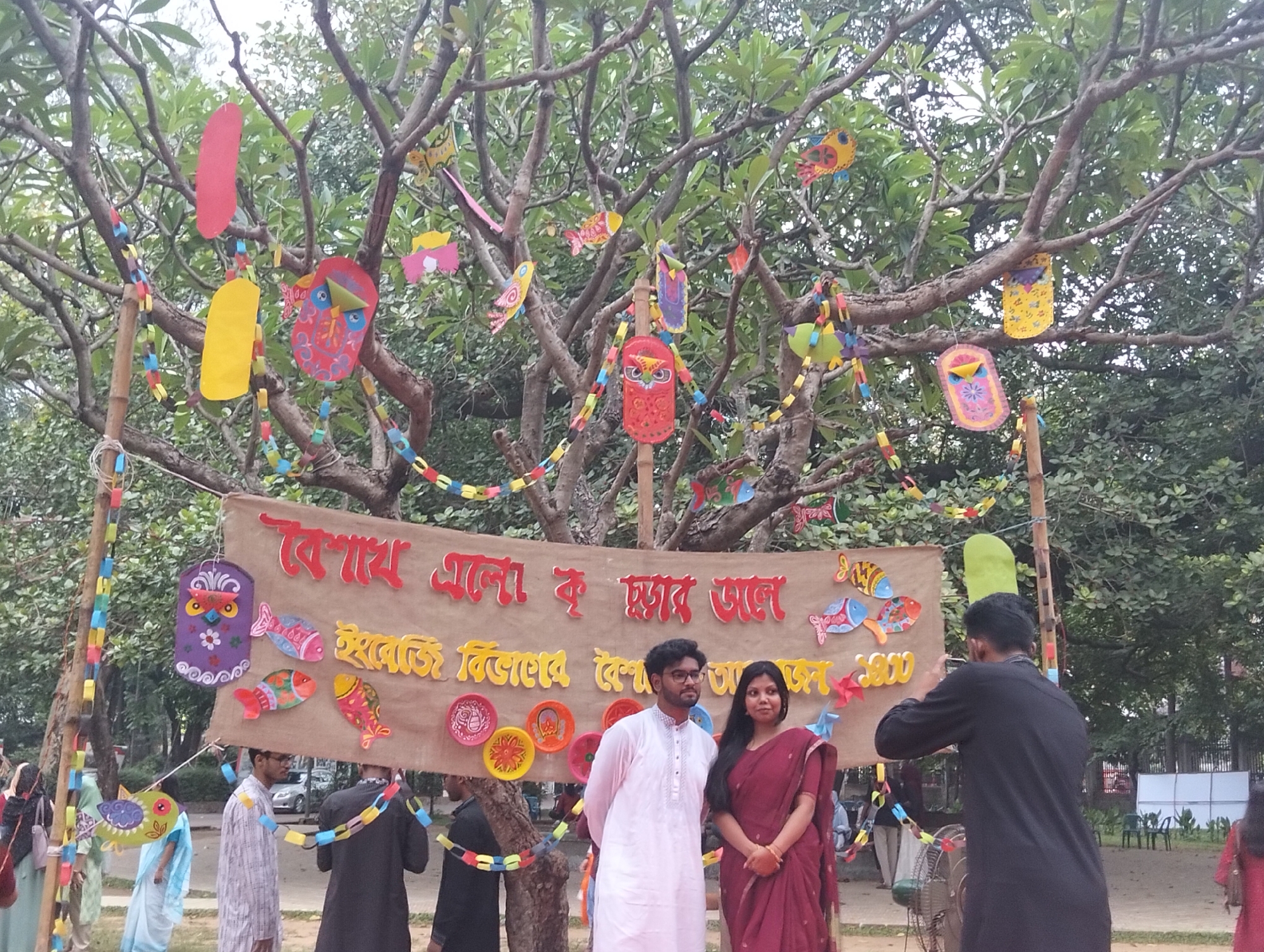 This image captures a vibrant outdoor festive event in Dhaka, Bangladesh, likely celebrating Pohela Boishakh, the Bengali New Year. In the foreground, a man and a woman are posing for a photograph. The man wears a white Punjabi (kurta) and the woman is dressed in a maroon saree. To their right, another man, wearing a black Punjabi, is actively taking their picture with a smartphone.
They stand in front of a large, brown fabric banner decorated with colorful cut-outs of fish and round, patterned plates. The banner prominently displays Bengali text. The top line reads "বৈশাখ এলো ক হাজার ভাল" which translates to "Boishakh has come, how good it is." Below this, another line of text is partially visible, starting with "ইবতেজী বিলবের বে".
The setting is beneath a large tree with lush green foliage. The tree branches and bamboo poles supporting the banner are extensively decorated with numerous handmade, colorful paper items, including fish, birds, and abstract shapes, as well as long chains made of small, interconnected paper rectangles. These decorations contribute significantly to the festive ambiance.
Other individuals are visible in the background, some partially obscured, suggesting a gathering. The natural light indicates it is daytime, possibly under an overcast sky, which provides soft, even illumination. A standing fan is partially visible near the photographer. The overall scene conveys a celebratory mood, rich in cultural expression and community engagement.