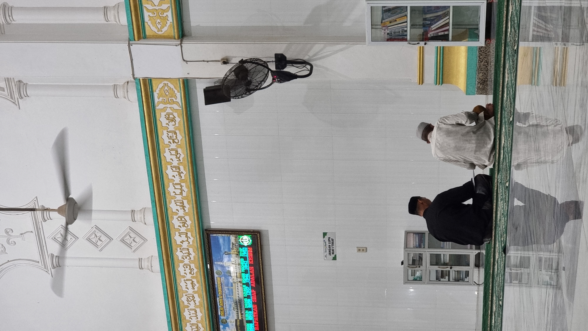 This is a vertical, medium shot of two men praying in a mosque in Lapang Timu, Indonesia. The man on the left is wearing a black shirt and pants and a black cap. He is facing away from the camera, with his hands clasped in front of him. The man on the right is wearing a striped shirt and pants and a light-colored cap. He is also facing away from the camera, with his hands resting on his lap.
The mosque interior is white with a tiled floor that reflects the light. Above the men, there is a decorative frieze with Arabic calligraphy in gold and green. A ceiling fan is visible in the upper portion of the image, and a wall-mounted fan is to the right. To the left of the praying men is a small white cabinet with glass doors, filled with books. To the right of the praying men is a white bookshelf filled with books. There is a digital display screen above the bookshelf, which shows an image of a mosque and text in Arabic and Indonesian. A small green sign is also visible on the wall, with Indonesian text. The overall impression is of a serene and devout atmosphere.