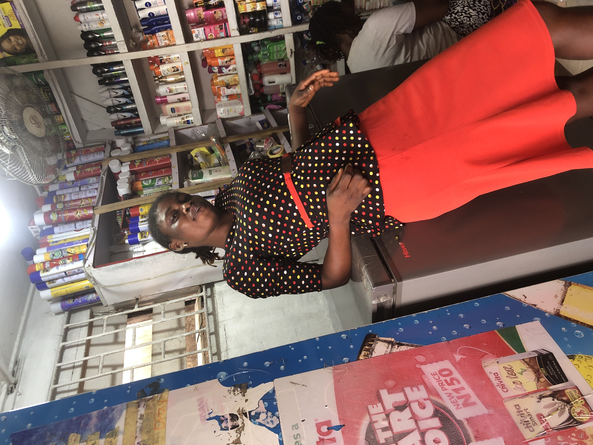A woman in a black, red, and yellow polka dot blouse and a red skirt leans against a refrigerator in what appears to be a small convenience store or market stall. The woman has dark skin and her hair is braided. Behind her, shelves are stocked with various products, including bottles of deodorant or hairspray, skincare products, and food items. A ceiling fan is visible above the shelves. To the left of the woman, a blue and red advertising poster is partially visible, featuring text and images of beverages. The overall environment suggests a busy retail setting. The image is shot from a slightly high angle.