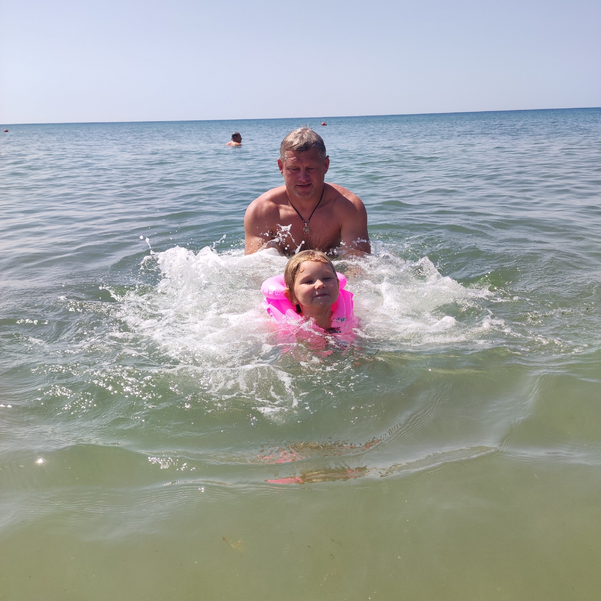 In this image, a man and a young girl are enjoying a day at the beach. The man, with light brown hair and a tanned complexion, is partially submerged in the water behind the girl. He is wearing a dark necklace. The girl, with blonde hair, is in the foreground and is wearing a bright pink life vest. She has a slight smile on her face, looking towards the camera.
They are in the ocean, with gentle waves creating splashes of water around them. The water is a murky green color, and sunlight sparkles on the surface. In the background, the vast expanse of the ocean meets a clear, pale blue sky, suggesting a sunny day. There are a couple of other figures in the distance, also in the water, indicating a public beach setting. The overall scene conveys a sense of family fun and enjoyment of a summer day. There is no visible text in the image.