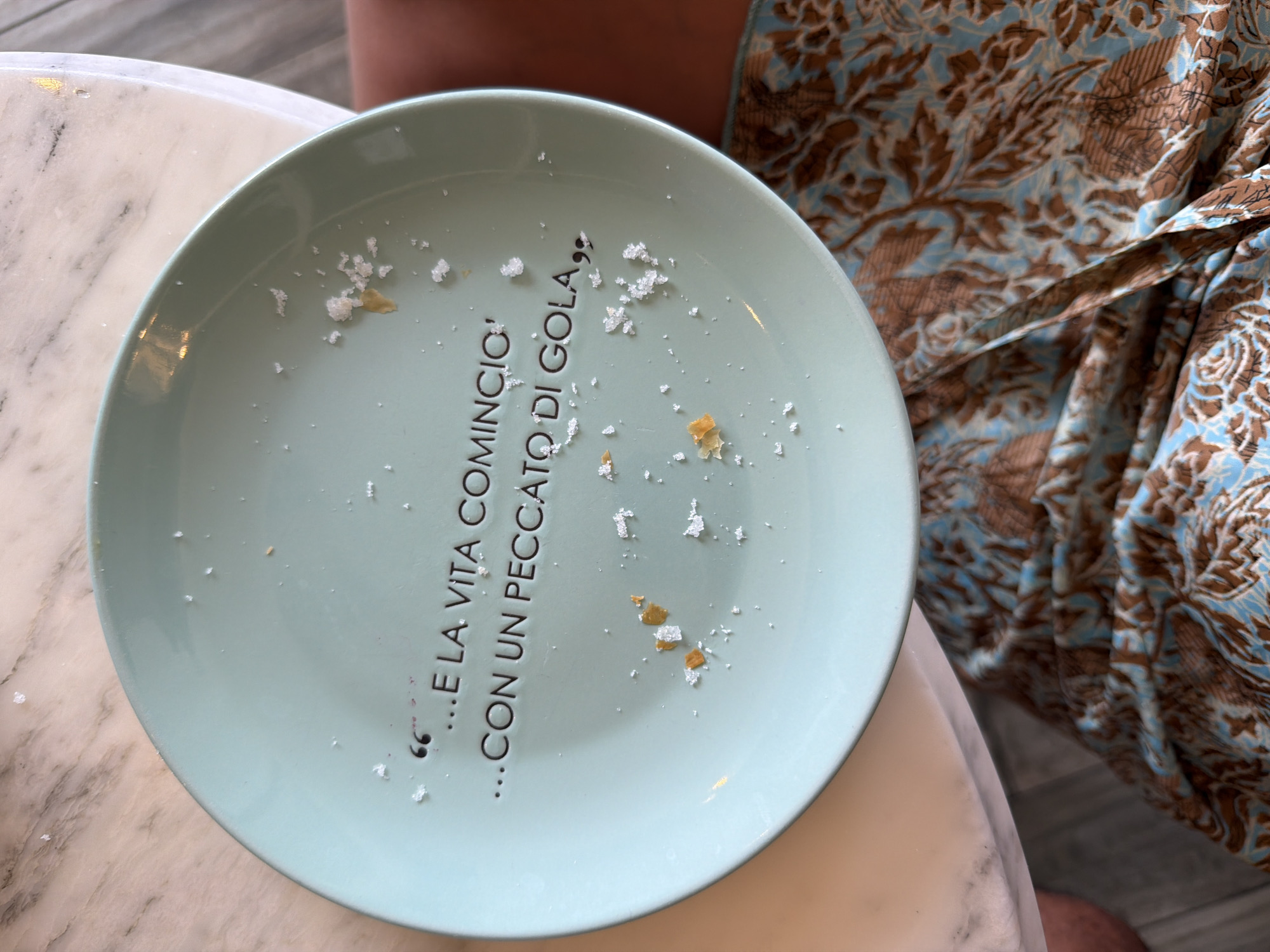 The image shows a pale green plate on a marble-topped table. The plate has black text in Italian that reads, "...E LA VITA COMINCIO"...CON UN PECCATO DI GOLA,, which translates to "...And life began"...with a sin of gluttony,,. There are crumbs and a few scattered white specks on the plate, suggesting it was recently used. A person's legs, clad in patterned fabric, are visible in the lower portion of the image, indicating the plate is part of a meal setting. The scene is an indoor setting, likely a restaurant or home dining area. The image does not provide any information about the city of Olbia, Italy.
