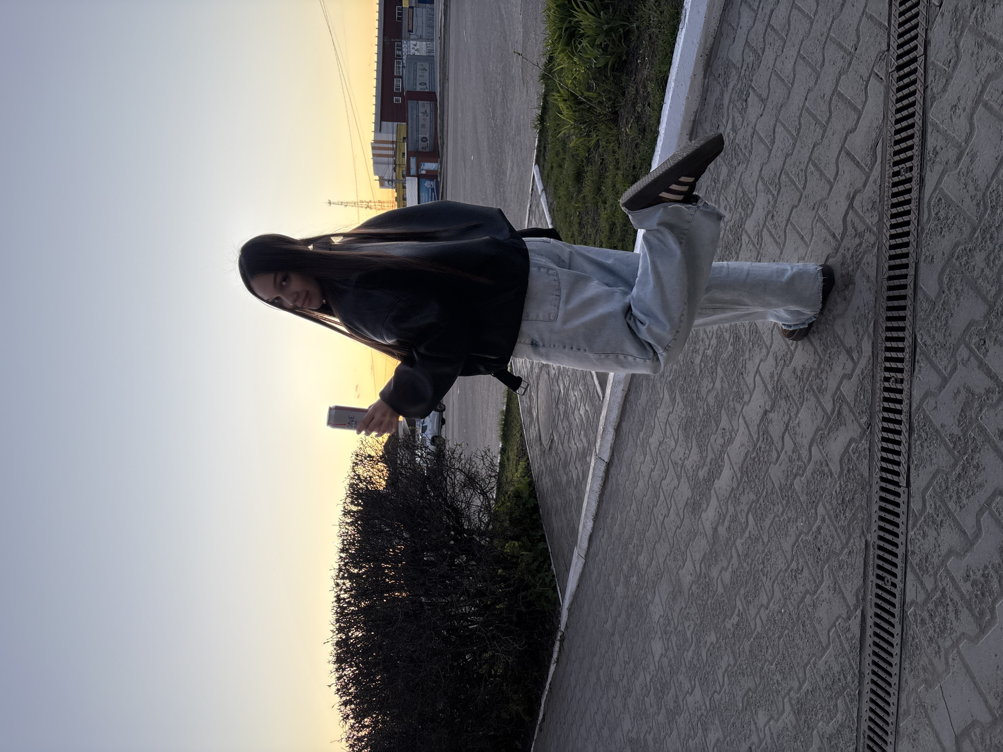 A young woman stands on a paved surface, likely a sidewalk, next to a drainage ditch. She is wearing light blue jeans, a black jacket, and Adidas shoes. In her hand, she holds a can of something. The background features a building complex, possibly industrial or commercial, and a partially obscured sunset or sunrise. The location is consistent with an urban environment, potentially Chaikovsky, Russia.