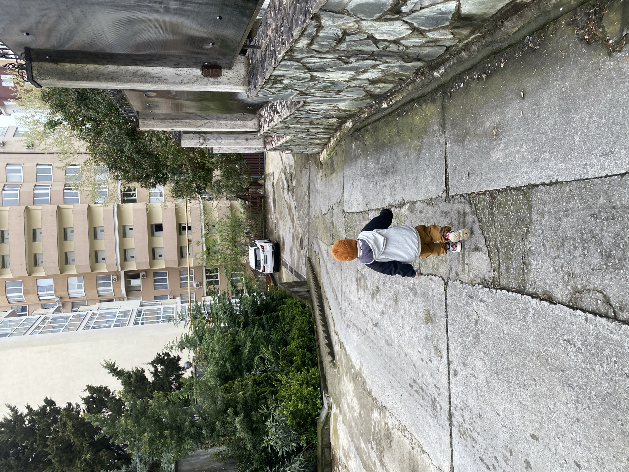 A child wearing an orange beanie, a white hoodie, brown pants, and mismatched sneakers is standing on a concrete walkway, seemingly looking down at something. The walkway is bordered on one side by a stone wall and on the other by a building and a green shrubbery. In the background, a white car is parked. The buildings suggest an urban environment, and based on the location identified, the setting is likely Sochi, Russia.