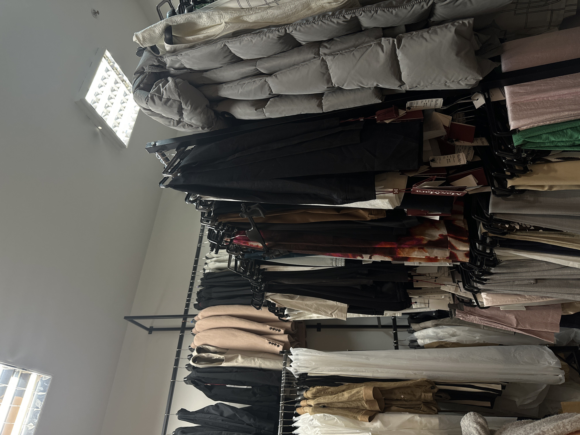 The image shows racks of clothing in a store, likely a high-end boutique, possibly in Sofia, Bulgaria. The clothing items include a puffy down jacket, suits, blazers, dresses, pants, and shirts. Many of the clothes have price tags. The racks are organized vertically along a wall, with natural light coming from a window near the top of the frame. The environment appears clean and well-lit.