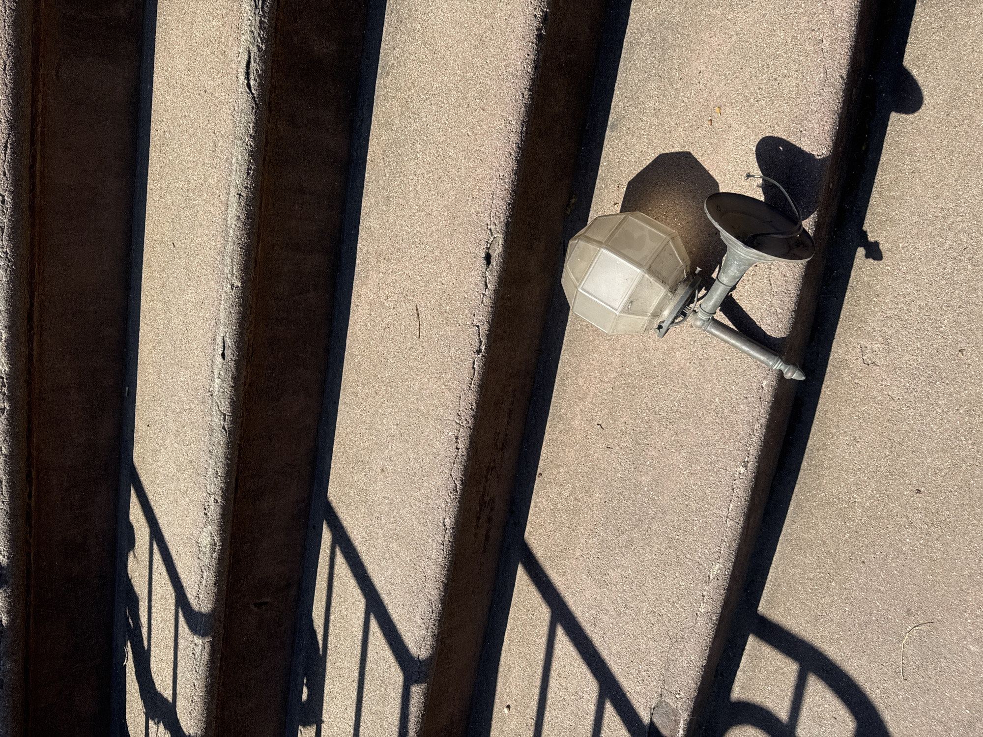 The image shows an outdoor staircase made of light grey concrete. Shadows cast by the railing run diagonally across the steps from the lower left to the upper right. In the upper right corner of the image lies an old, broken outdoor light fixture. It has a spherical, multi-faceted glass shade, a conical metal shade, and a connecting metal arm. The overall scene appears to be a city street in New York City, USA, based on the architectural style.