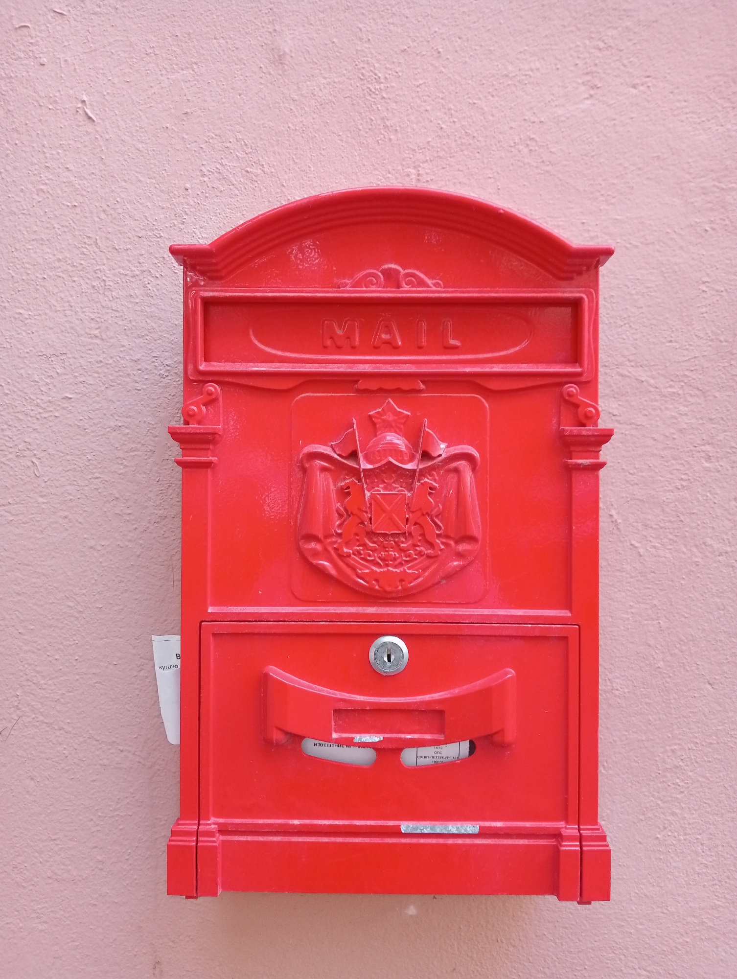 A bright red, ornate mailbox is mounted on a textured pink wall. The mailbox features a curved top with ridged details and the word "MAIL" embossed in raised lettering. Below the "MAIL" inscription is a decorative crest with two lions and a shield. A small compartment with a lock is located below the crest, and a red handle is positioned above it. A piece of white paper with Russian text is visible peeking out from the left side of the mailbox. The wall appears to be exterior stucco. The context of Saint Petersburg, Russia is inferred by the Russian text on the paper.