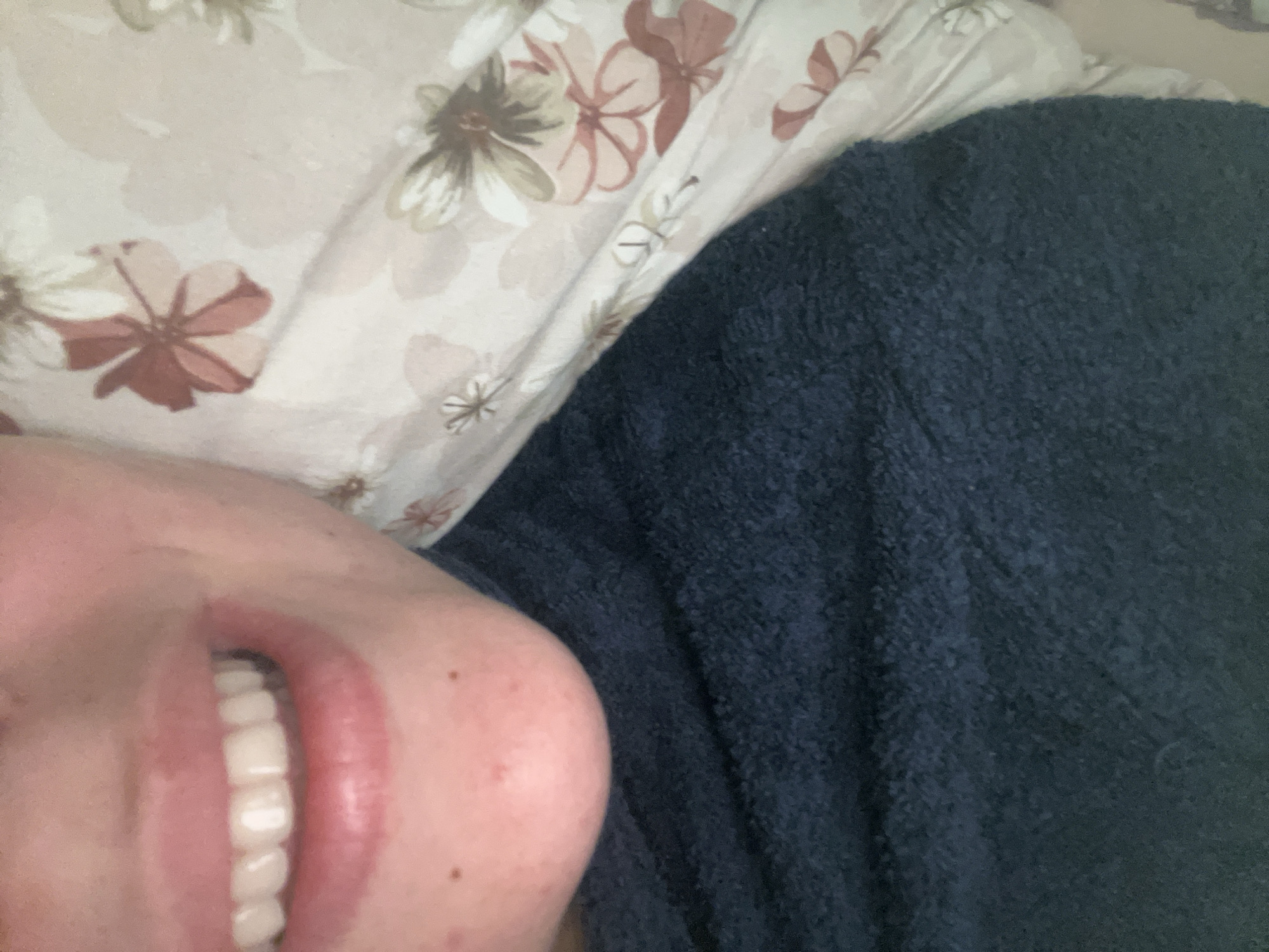 The image shows a close-up, sideways view of a person's face and a dark blue textured blanket. The person's lips are slightly parted, revealing white teeth. Their skin is visible on the left side of the frame, with a few small dark spots. The person appears to be resting against a white pillow with a floral pattern in muted tones of pink, beige, and green. The activity taking place is unclear, but the positioning suggests the person might be lying down. The lighting is soft, and no specific time of day is discernible. No text is visible in the image. The location context is Plovdiv, Bulgaria.