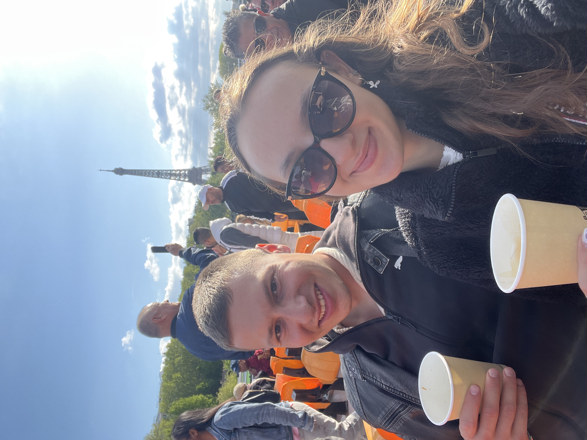 This photograph captures two smiling individuals in the foreground, a young woman and a young man, enjoying a sunny day outdoors in Paris, France, with the iconic Eiffel Tower prominently visible in the background.
The young woman, positioned slightly above and behind the man, has long, wavy brown hair and is wearing black sunglasses, a dark, textured jacket, and a distinctive butterfly-shaped earring on her left ear. Her smile is directed at the camera, and reflections in her sunglasses show hints of the surrounding environment. Beside her, the young man with short, light hair also smiles warmly at the camera. He is dressed in a dark jacket over a lighter top. Both individuals are holding light-colored paper cups, suggesting they are enjoying a drink.
The setting appears to be a public viewing area or an event, with several other people visible in the blurred middle ground and background, along with rows of orange, stadium-style seats. The weather is bright and clear, with a vivid blue sky dotted with white clouds. The Eiffel Tower stands tall in the far background, confirming the location as Paris. The overall atmosphere is cheerful and relaxed, indicating a pleasant experience. No discernible text is visible on any objects or clothing in the image.