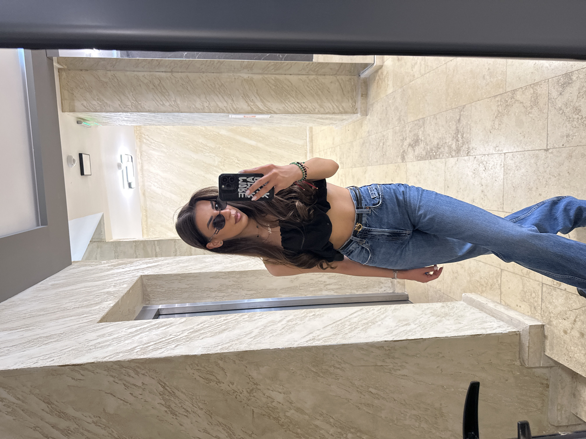 A young woman is taking a mirror selfie in a building hallway. She is wearing a black crop top, blue jeans, and sunglasses. The hallway has textured walls and a tiled floor. An elevator door is visible to her left. The scene appears to be in the city of Sofia, Bulgaria.