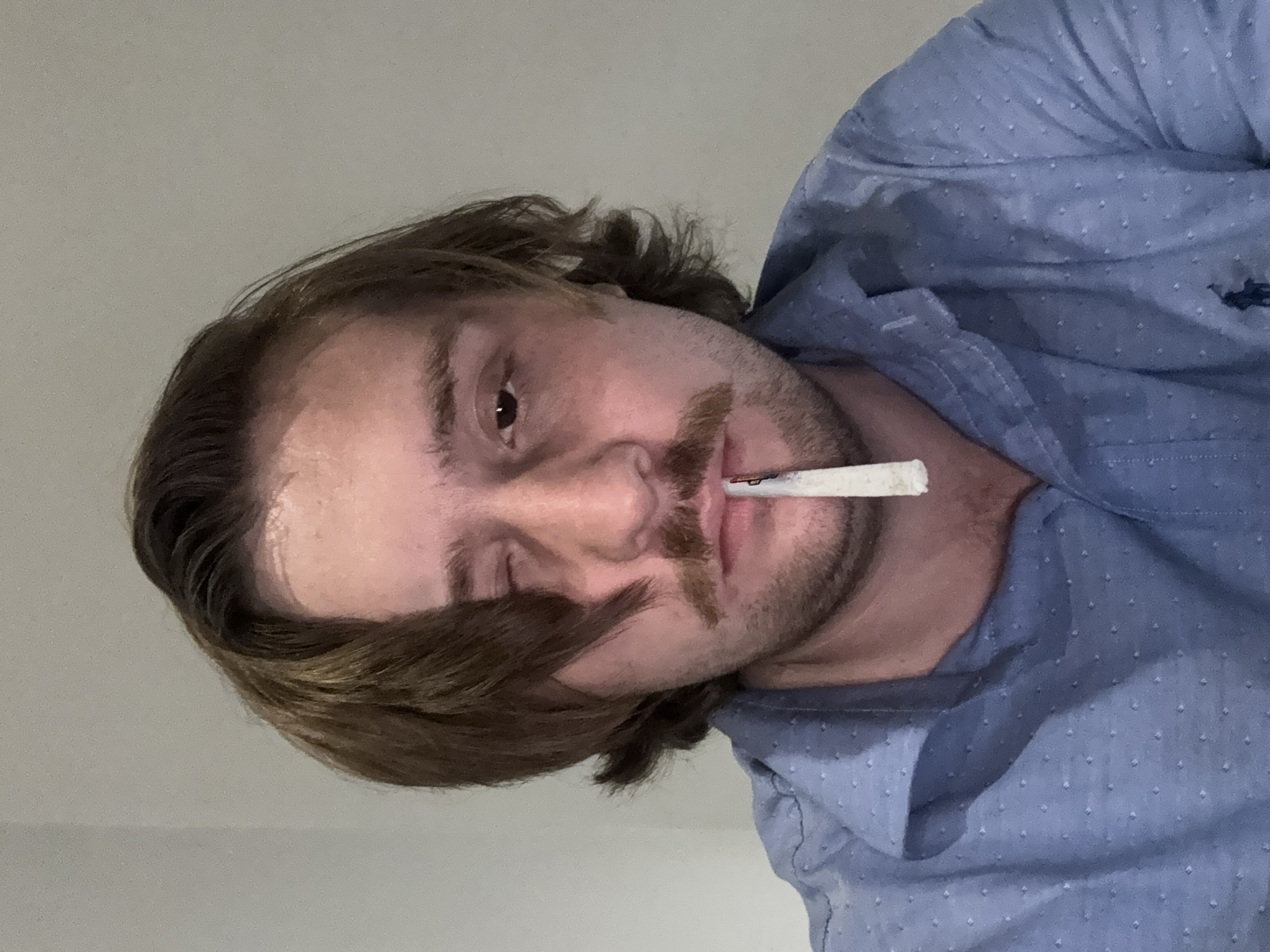 A person with long brown hair and a mustache holds a rolled cigarette in their mouth. They wear a light blue button-down shirt with small white dots. The background is a plain white wall. There is no evidence in the image to determine if the image was taken in Huixquilucan de Degollado, Mexico.
