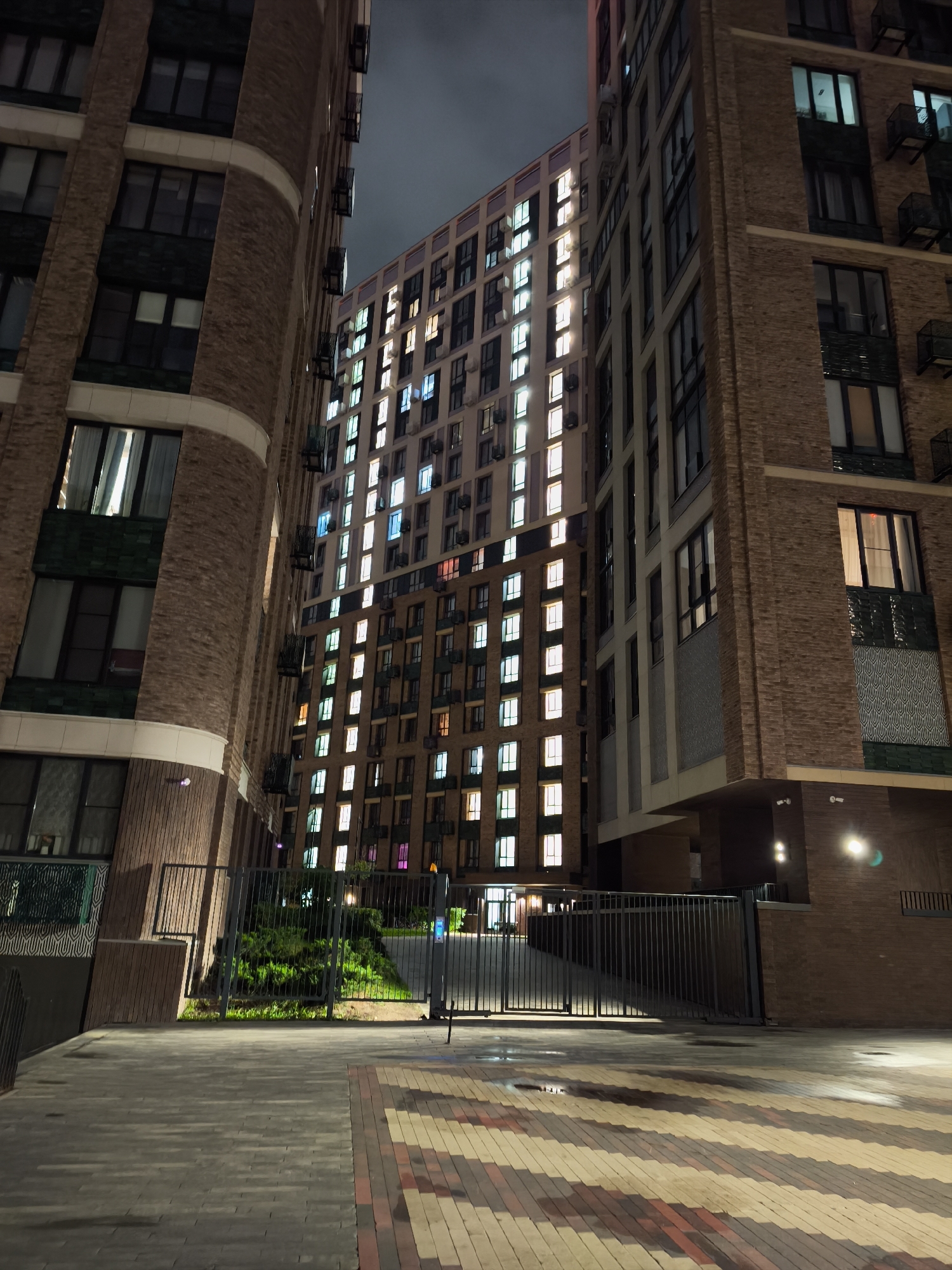 The image depicts a view of modern residential buildings at night in Moscow, Russia. The buildings are constructed of red brick with symmetrical window arrangements. Most windows are illuminated, suggesting occupancy. A black metal gate is in the foreground, guarding a pathway leading up a gentle slope covered with greenery. The paving stones are a mix of gray and red bricks laid in a pattern. The sky is dark, indicating nighttime. No people are visible in the frame. The lighting suggests a well-lit urban environment.