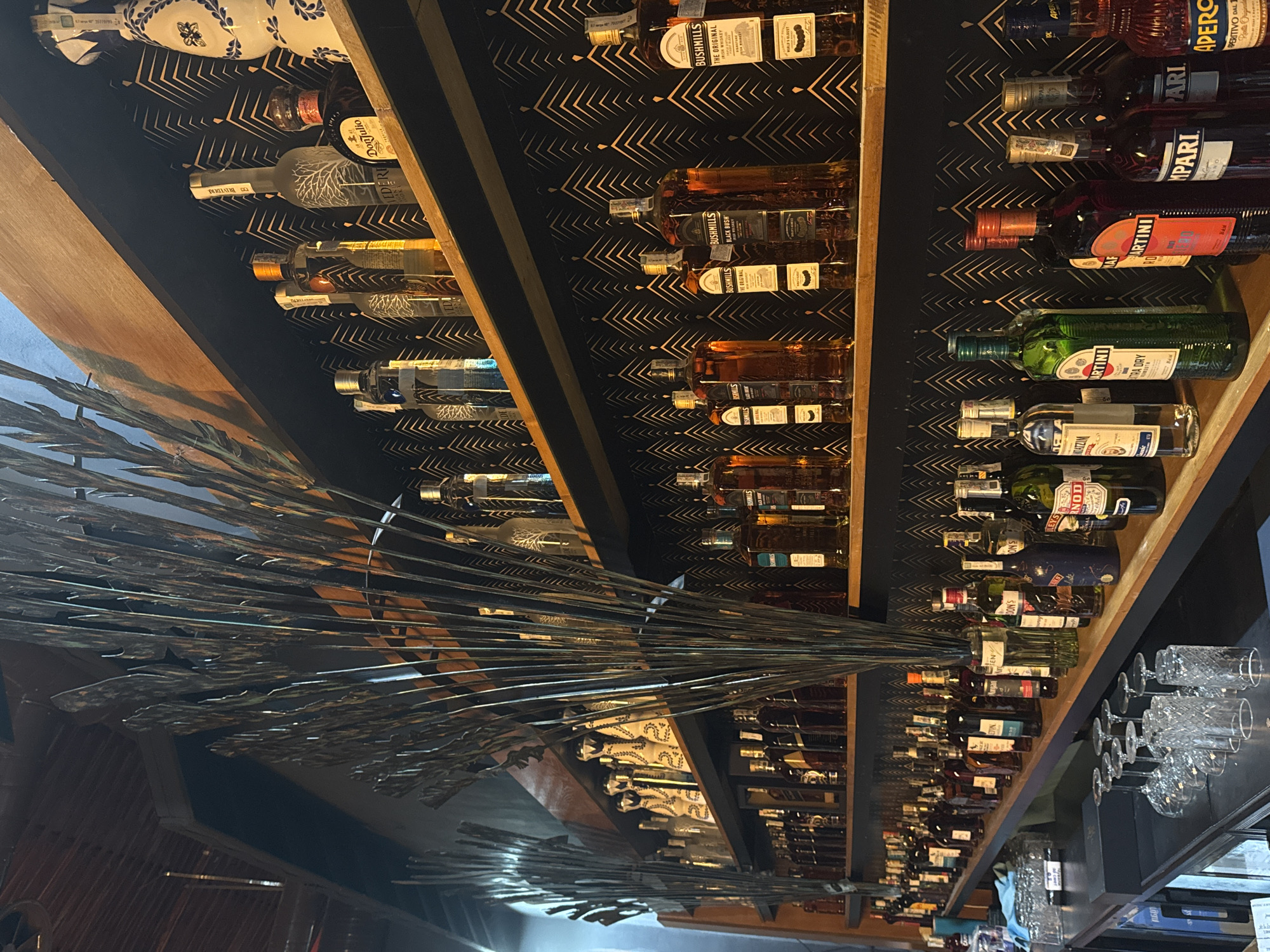This media file captures an interior scene, likely a bar or a well-stocked liquor section, in Tsarevo, Bulgaria. The primary focus is on multiple shelves filled with a variety of alcohol bottles. The shelves are made of wood and extend diagonally upwards from left to right, creating a dynamic perspective.
The background behind the bottles features a dark surface adorned with a repeating, geometric pattern of light-colored, angular lines. In the foreground, spanning from the lower left towards the upper right, is a prominent, abstract decorative element composed of numerous thin, metallic or dark-colored stalks or reeds, fanning out like foliage.
Numerous liquor bottles are visible, showcasing various well-known brands. Recognizable brands include:
- Tequila: Don Julio (identifiable by its distinct blue and white patterned bottle).
- Vodka: Belvedere (with its characteristic frosted tree design).
- Whiskey: Bushmills (several bottles, including "The Original" and "Black Bush").
- Vermouth/Aperitif: Martini ("Extra Dry" and "Rosso" variants), Campari, and Aperol (an orange-hued bottle with "APERITIVO DAL" visible).
Other partially visible labels include "DON'S," "LEYS," "NOD," "ATZIM," and "BAN."
On the far right, a bar counter is visible, topped with a stack of ornate, cut-glass stemmed glasses, suggesting the preparation or serving of drinks. No people are visible in the image, indicating a static display within the establishment. The artificial lighting suggests an indoor setting.