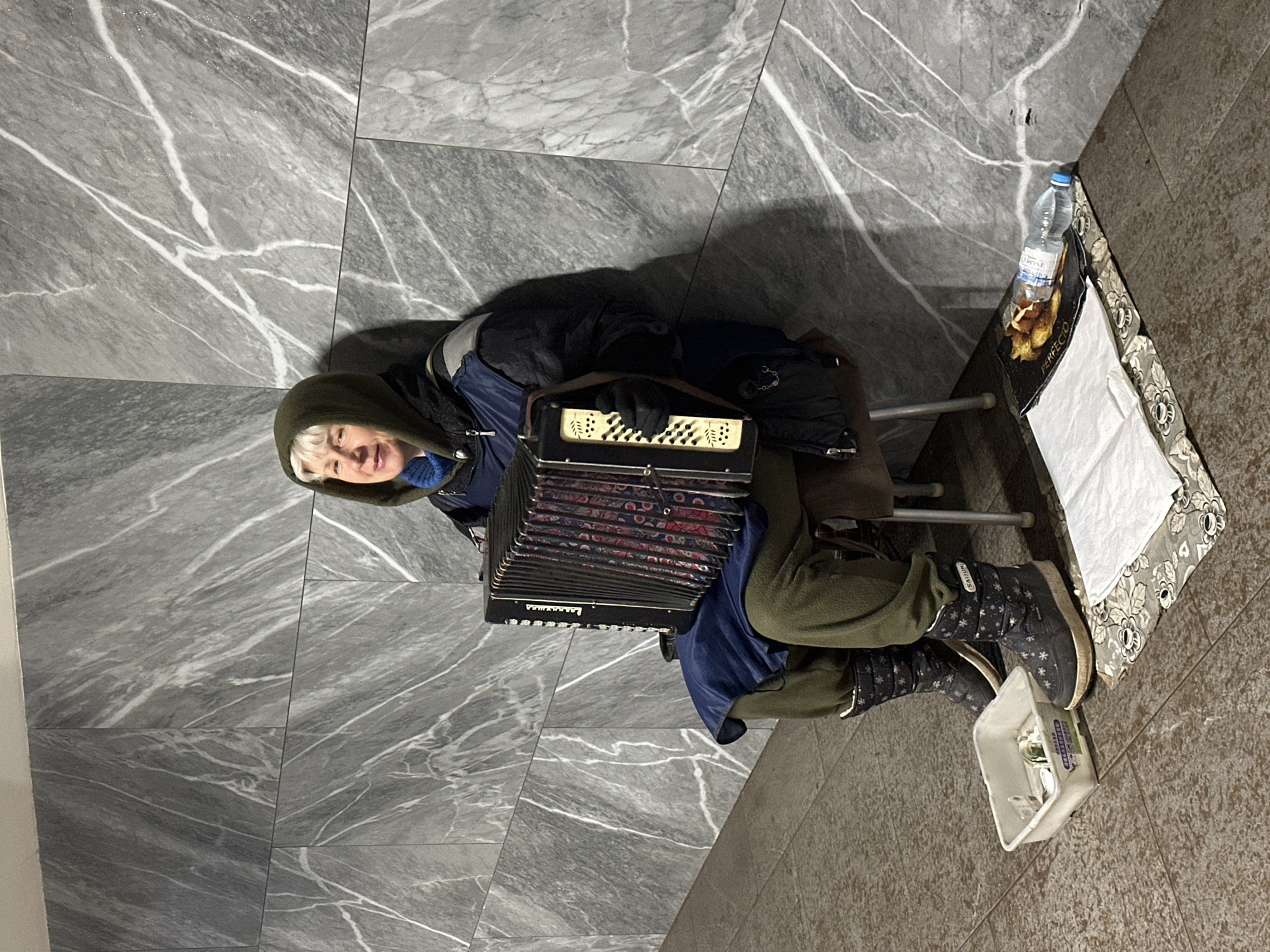 A woman wearing an olive-green hooded coat and dark winter boots sits on a stool in a subway station, playing an accordion. She has short, light-blonde hair and is smiling. Next to her, on a floral-patterned cloth, is a water bottle and a tray with food. A white plastic container filled with money sits near her boots. The wall behind her is covered in gray marble tiles with white veining. The location is likely a subway station in Nizhny Novgorod, Russia.