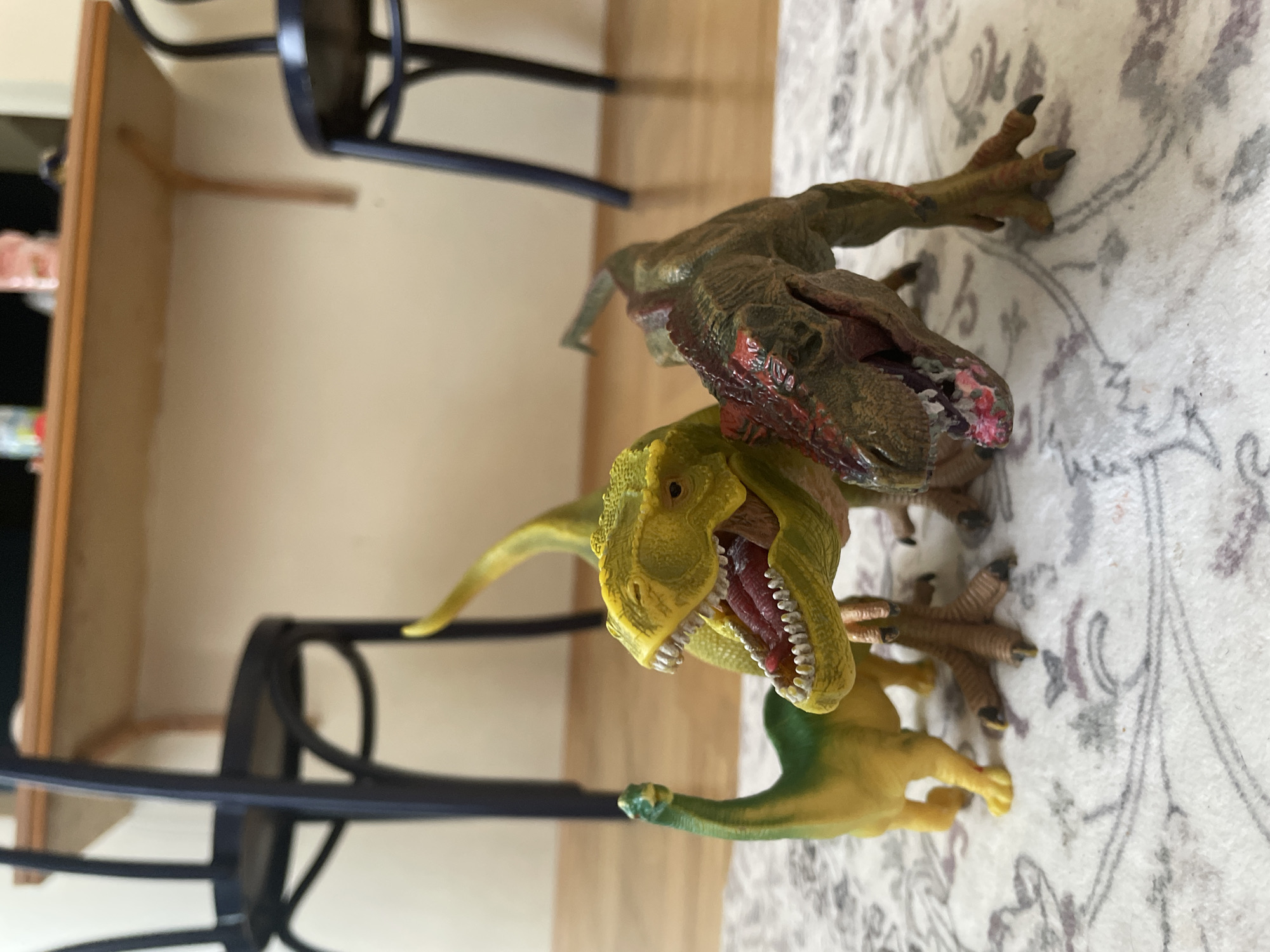 In a domestic setting, possibly an indoor area with a wooden floor and a patterned rug, three toy dinosaurs are arranged on the rug. In the foreground, a green dinosaur with a long neck stands to the left. To its right, a larger yellow dinosaur with its mouth open is positioned prominently. Partially behind and to the right of the yellow dinosaur is a brown and red dinosaur, also with its mouth open. The background shows the legs and seats of two dark chairs and a wooden table. The overall lighting suggests an indoor environment, possibly with natural light contributing.