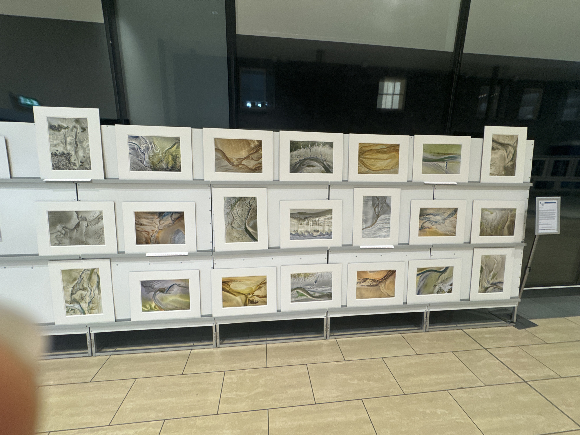 The image depicts an art exhibition featuring numerous framed abstract landscape paintings displayed on white shelving units. The paintings exhibit a range of earthy tones and fluid, organic patterns, suggesting the use of mediums like watercolor or acrylics. Some artworks appear to be monochrome, while others incorporate subtle color variations. The exhibition is set against a backdrop of large windows that reflect the interior lighting and the exterior environment, hinting at a contemporary building or gallery space. A sign with text is visible on the right side of the display, likely providing information about the exhibition or individual artworks. The flooring consists of large, rectangular beige tiles. The overall atmosphere is calm and organized, typical of an art display. The location in the city of Kilkenny, Ireland, is not explicitly evident from the visual information presented.