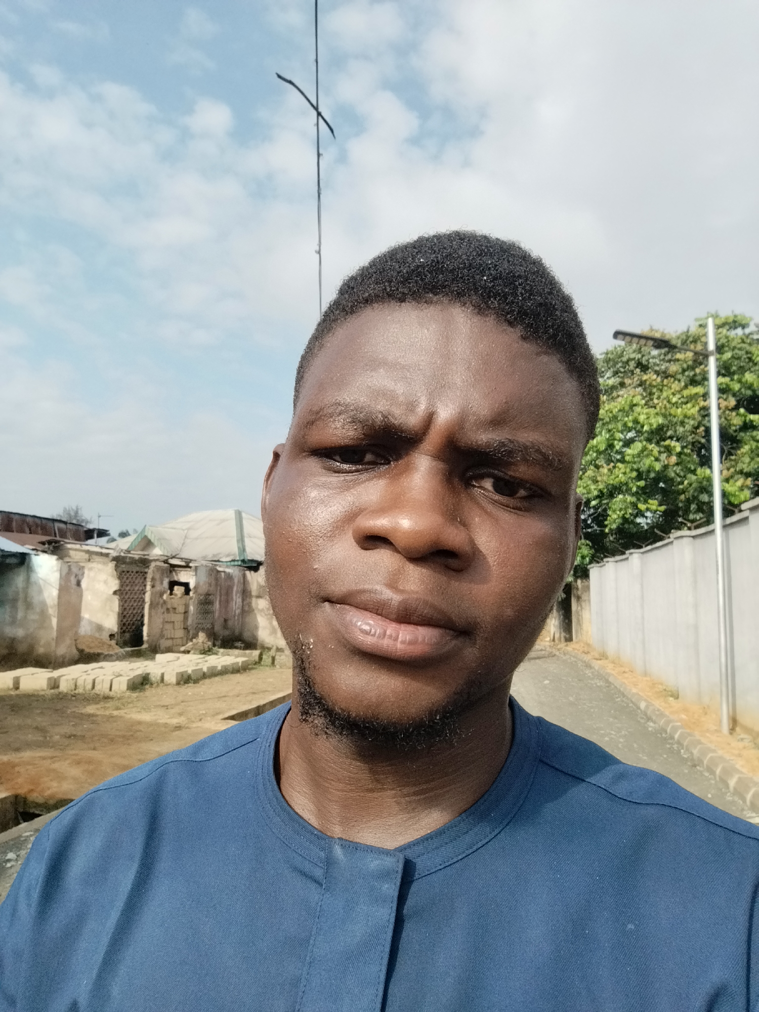 A medium, close-up shot of a dark-skinned man with short hair and a short beard, wearing a blue shirt. He is looking directly at the camera with a slight frown.
Behind him, there is a street scene in what appears to be Ikot Mbonde, Nigeria. To the left, there is a dilapidated building. To the right, there is a light gray concrete wall and a street with a curb. A street light is visible beyond the wall. The sky is pale blue with scattered white clouds.