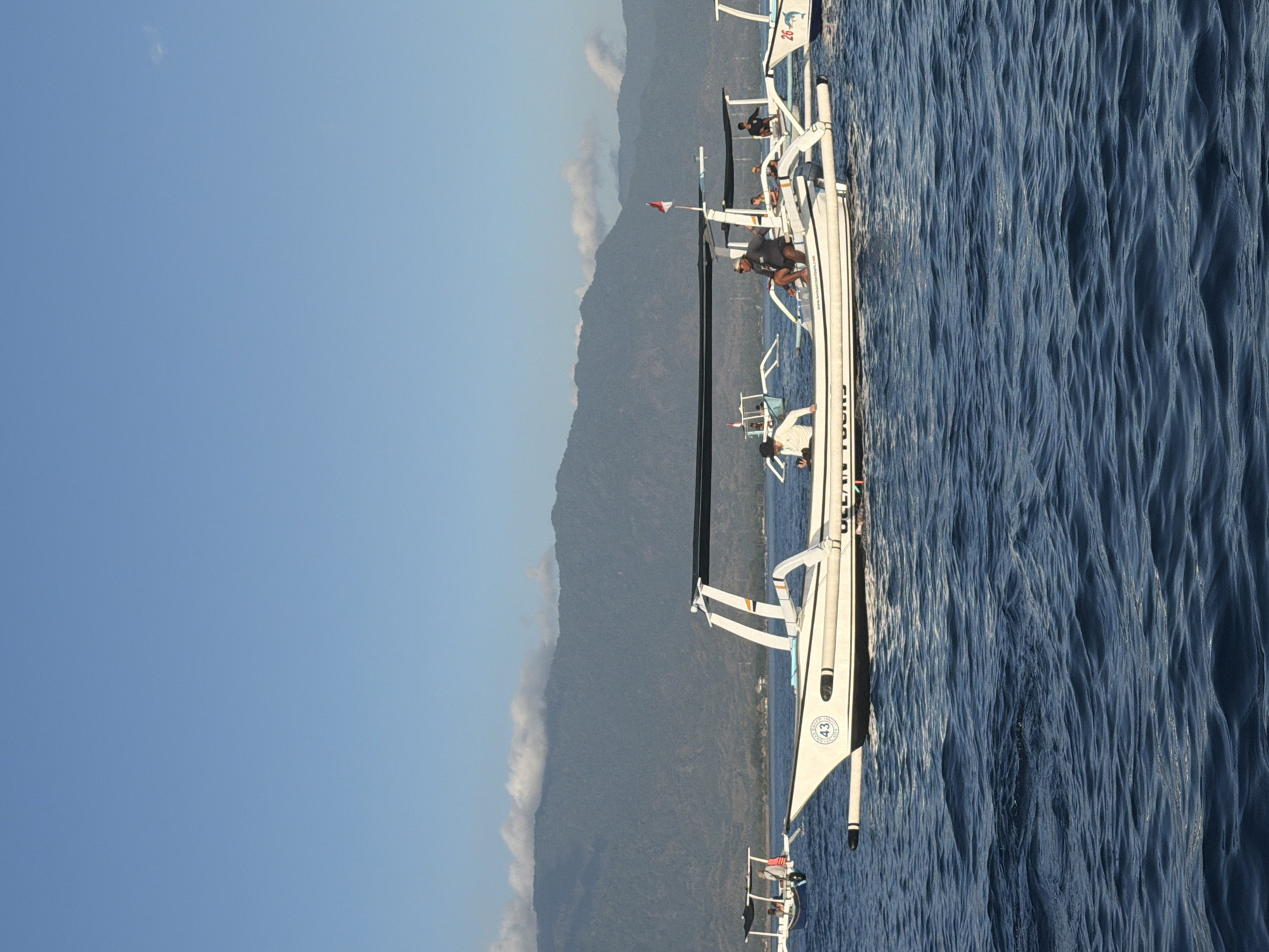The image depicts a scenic view of several white, outrigger boats on the water, with a lush, mountainous landscape in the background under a clear blue sky. In the foreground, a prominent white boat with a black canopy is positioned horizontally. This boat has the text "OCEAN TOURS" and a website address visible on its side. A circular emblem with the number "43" is also present on the hull. Several people are visible on this boat, some sitting and others standing, appearing to be passengers or crew members.
Further in the background, another white outrigger boat with a similar canopy is also visible, with people onboard. To the far left, a smaller blue and white boat is also present with passengers. The water is a deep blue with visible ripples and waves, suggesting a moderately active sea. The mountainous terrain in the background is densely covered with vegetation, and a line of white clouds stretches across the mountain range. The overall impression is that of a tour or excursion setting in a tropical coastal region. Given the context of "Ocean Tours" and the distinctive boat style, the location is likely a coastal area in Indonesia, possibly a popular tourist destination known for boat tours.