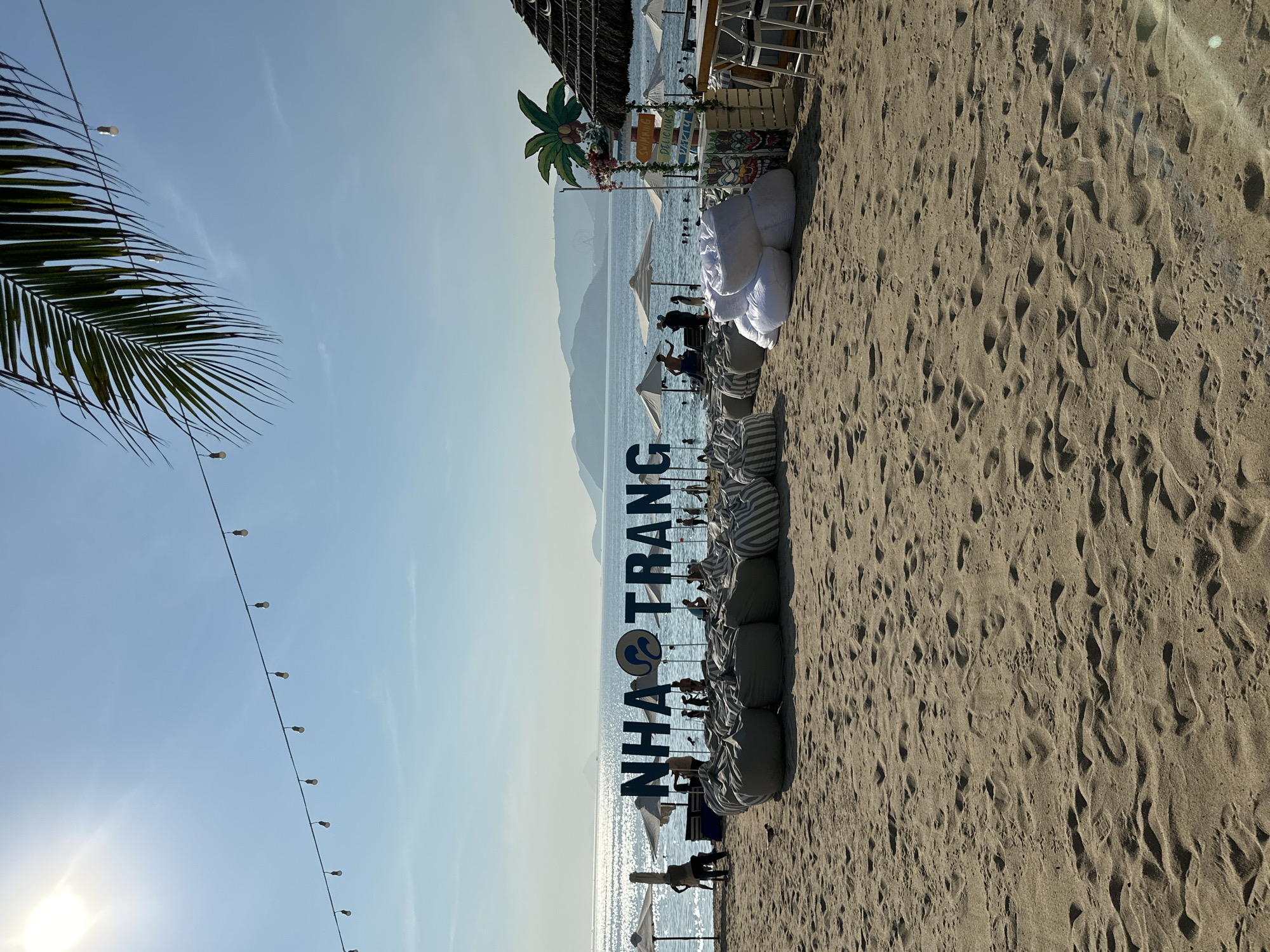 This image depicts a sunny day on a beach in Nha Trang, Vietnam. In the foreground, there are several large, plush beanbag chairs arranged on the sandy shore. Behind the beanbags, a large blue sign reads "NHA TRANG" with a circular logo in the center. Further back, people are seen relaxing and swimming in the ocean. Umbrellas are spread out on the beach, providing shade. The background features a hazy mountainous coastline. The atmosphere is that of a relaxed beach resort or a beachside establishment.