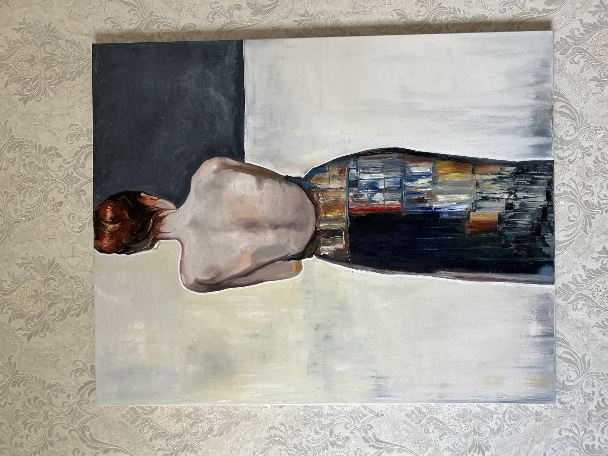 The image shows a painting of a woman from behind, with her back bare. She is wearing a dark skirt with a colorful, textured pattern around the waist. The background of the painting is divided into sections of light yellow, dark gray, and white. The painting is hung on a wall with a textured, damask-patterned wallpaper. The photograph is taken from a slightly angled perspective, looking up at the painting. There is no indication of the location being Moscow, Russia, or any other specific city.