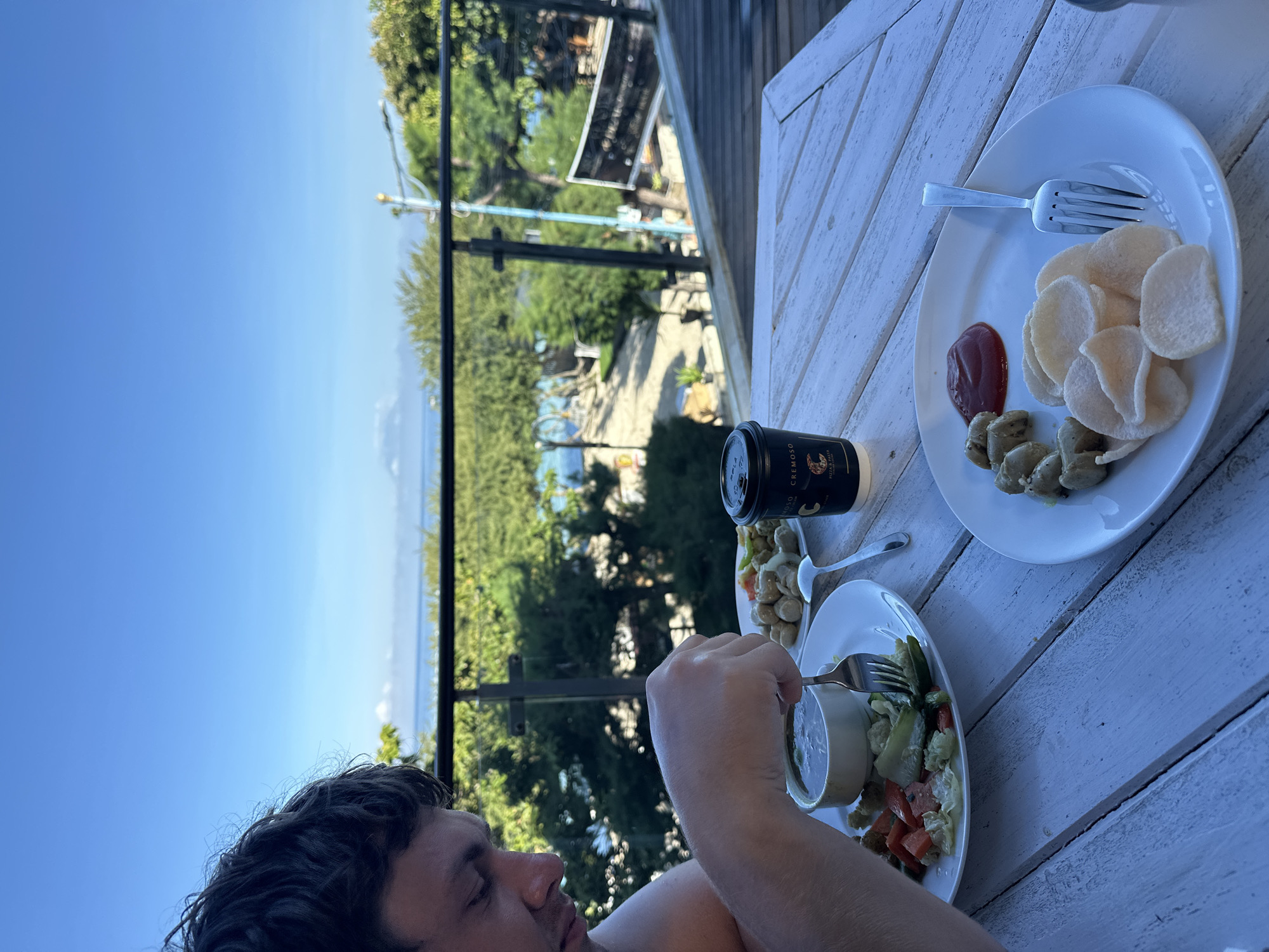 A man is seen from the torso up, eating food on a white wooden table on a balcony overlooking a view. The sky is blue, and the ocean is visible in the distance, along with green vegetation. The table has two white plates. One plate has what appears to be fried crackers, ketchup, and sausages. The other plate has a salad with a small bowl of dip. The man is using a fork to eat. A black coffee cup is also on the table. The city is Unknown, Indonesia.