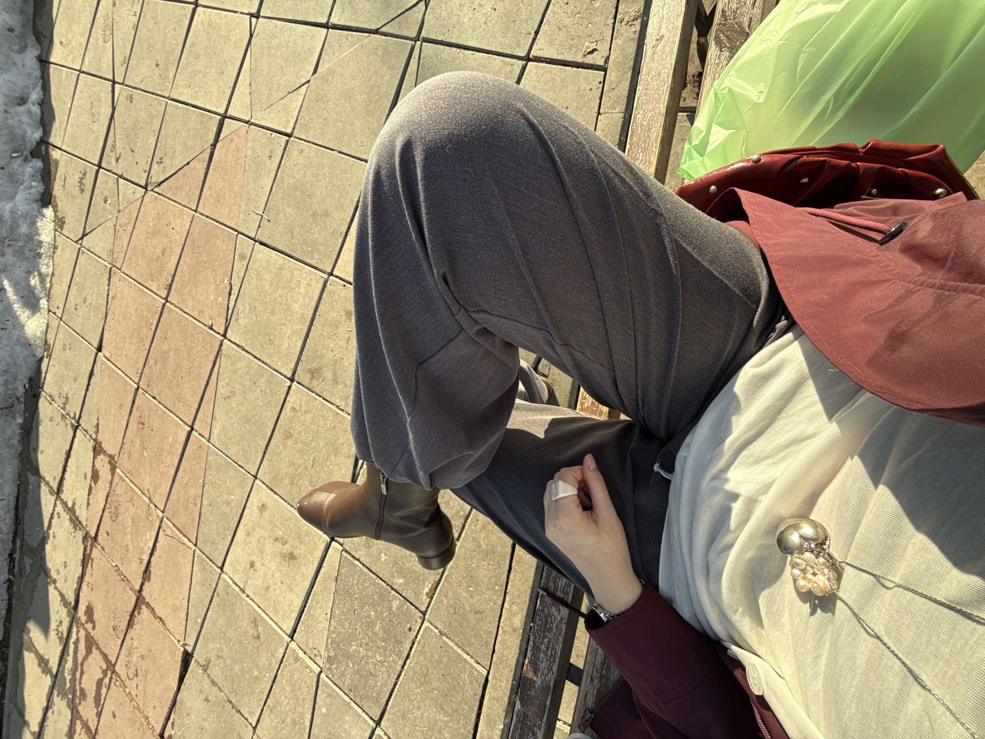 The image depicts a person sitting on a wooden bench outdoors. The person is wearing gray pants and brown boots. A maroon jacket and a green plastic bag are placed on the bench next to the person. The person's left hand is visible, adorned with a white ring. A silver necklace with pearl accents is visible on their white shirt. The ground is paved with square tiles in varying shades of beige and pink, suggesting a public walkway or park. A patch of snow can be seen at the edge of the walkway. The photograph's location is not discernible from the image.