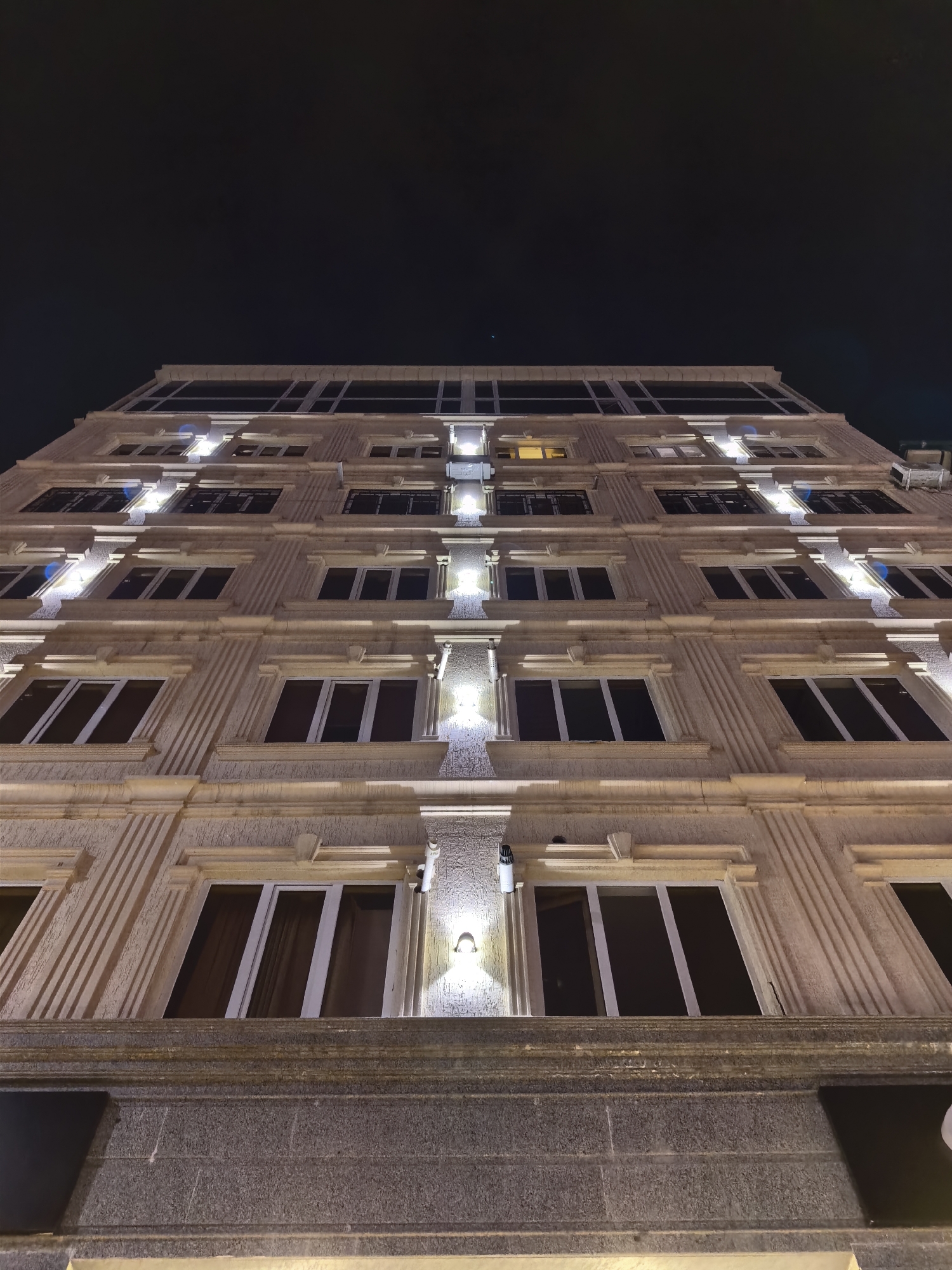 The image shows a low-angle, close-up view of the exterior of a modern building at night. The building has a beige facade with decorative architectural elements, including vertical fluting and horizontal cornices. Numerous windows with dark frames and reflective glass are visible, arranged in rows on multiple floors. Several exterior lights illuminate the facade, casting a warm glow on the textured surface and highlighting the architectural details. The sky above is dark and appears clear, with no stars or moon visible. The building's foundation is made of dark, rough-textured stone. The perspective emphasizes the height and grandeur of the structure. This building is located in Fergana City, Uzbekistan.